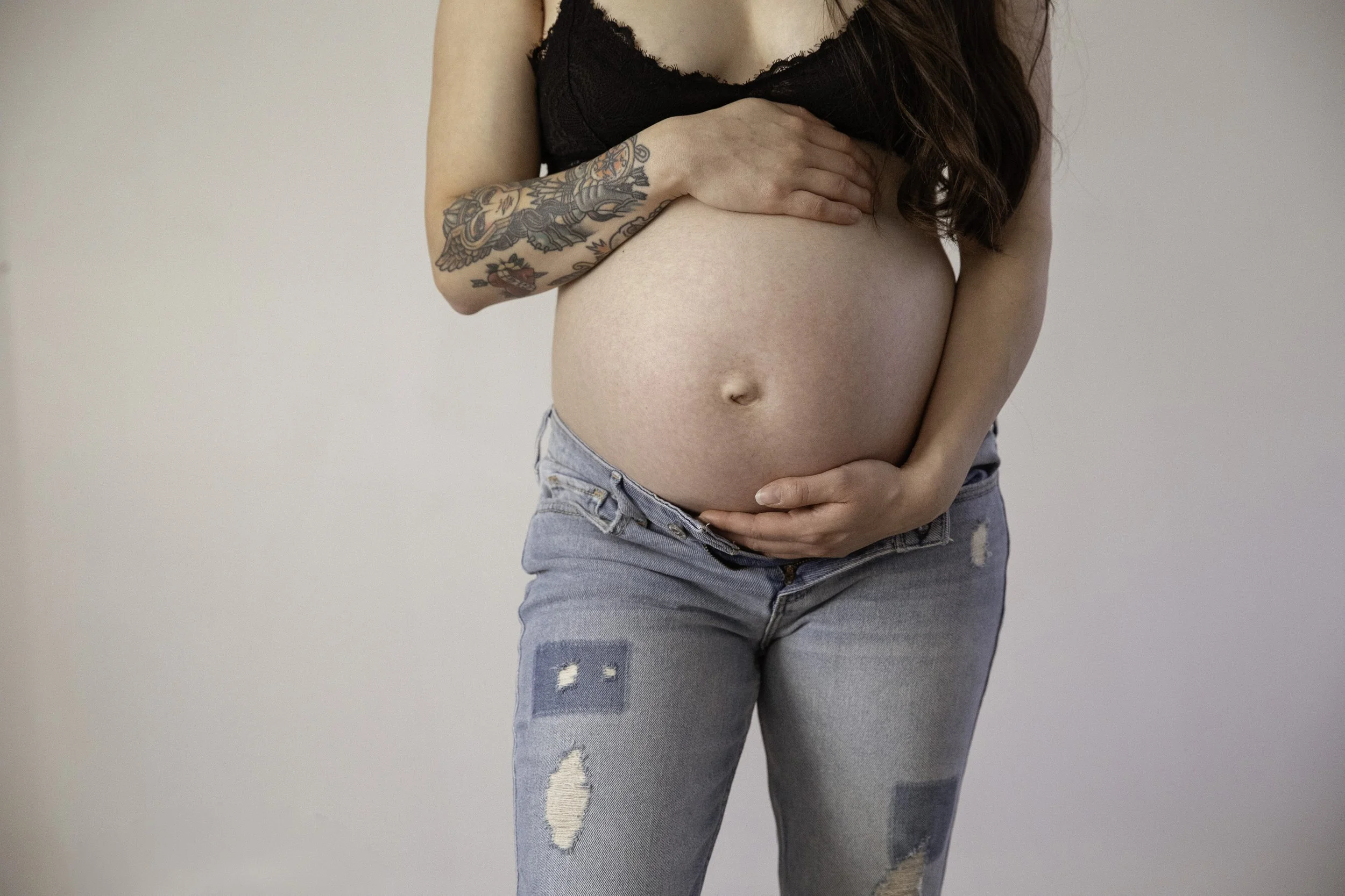 A woman with long brown hair wearing a black lace crop top and light blue ripped jeans, holding her pregnant belly with one hand and her other hand on her stomach, against a plain light gray background.