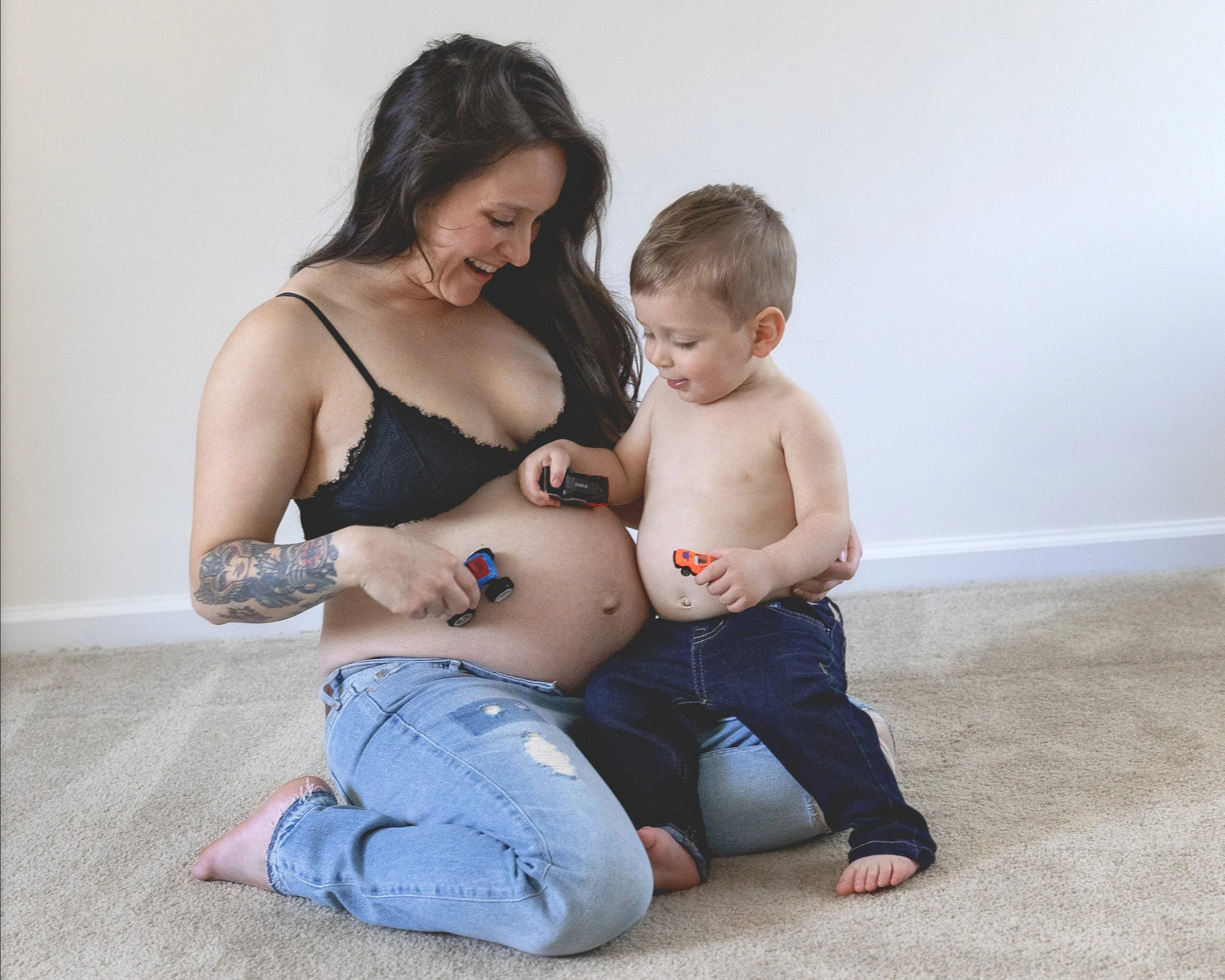 A woman with long dark hair and tattoos on her arm, smiling while holding a daughter with short blonde hair, both shirtless and playing with small toy cars on a beige carpeted floor against a plain light-colored wall.