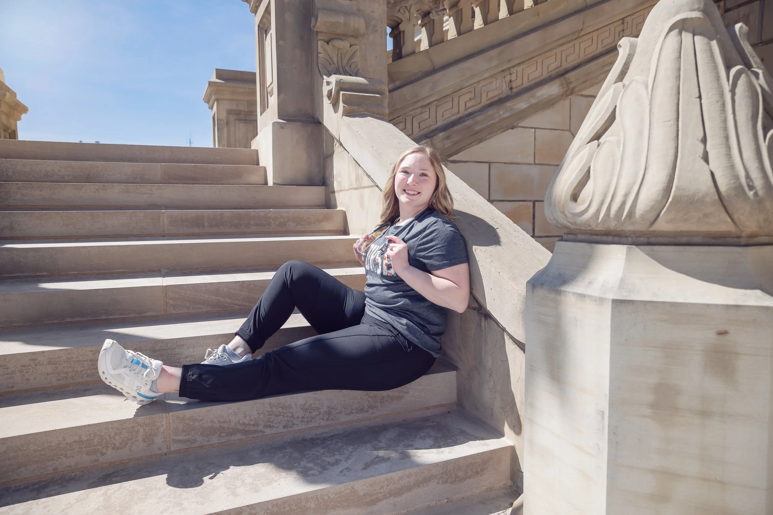 Young woman sitting on stone steps outside in front of a historic building, smiling, holding sunglasses, wearing a gray t-shirt and black pants, bright sunny day.