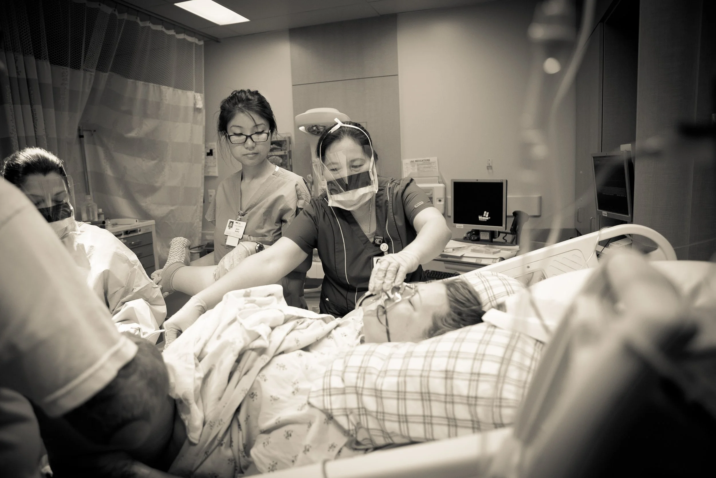 Healthcare professionals in scrubs and masks perform a medical procedure in a hospital room, with one patient lying on a bed surrounded by medical equipment.