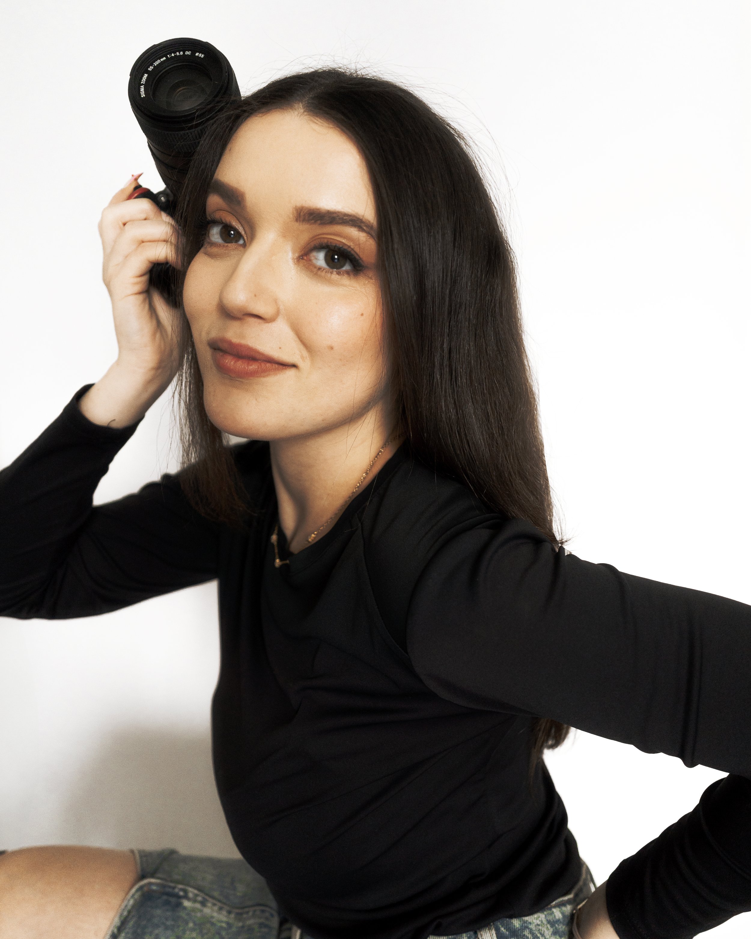 Femme tenant un appareil photo à côté de son visage, portant un haut noir, fond blanc.