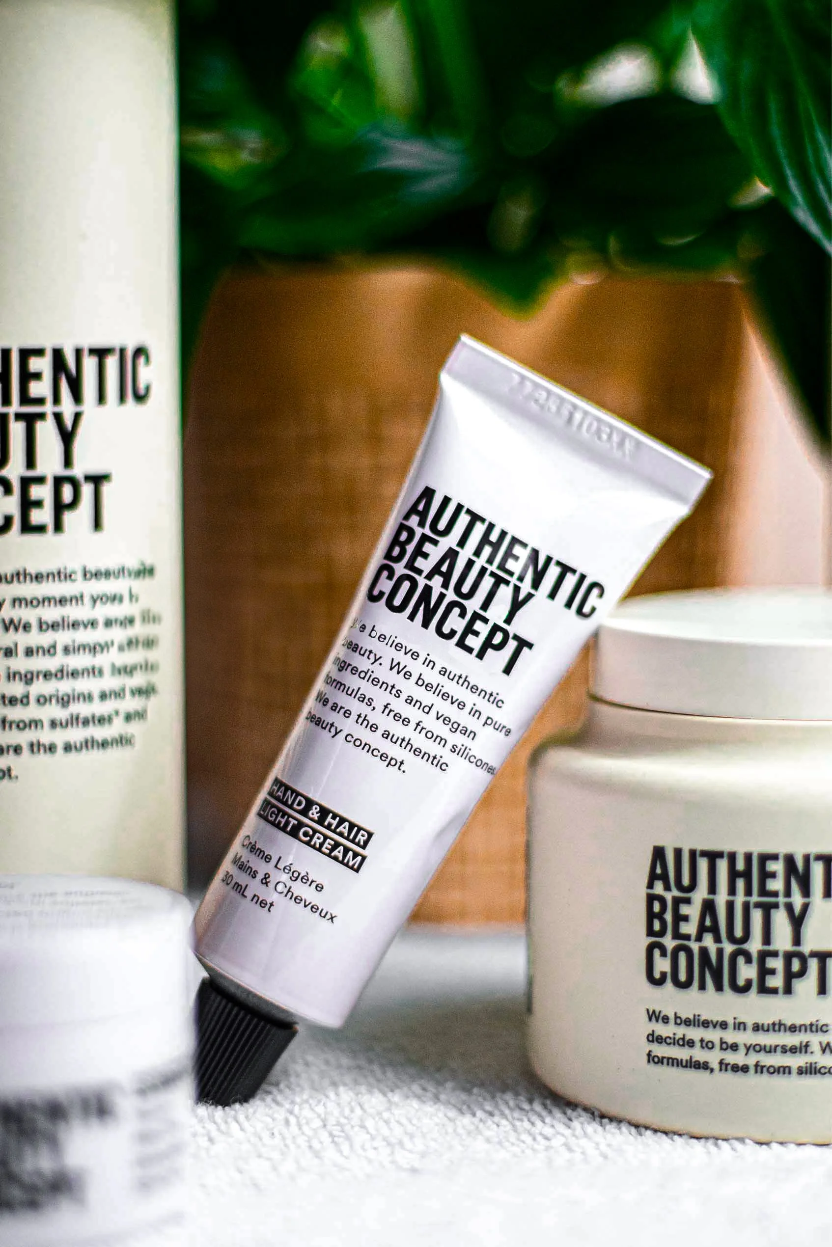 Produits Authentic Beauty Concept, y compris une crème pour les mains et les cheveux, avec des emballages minimalistes sur un fond végétal.