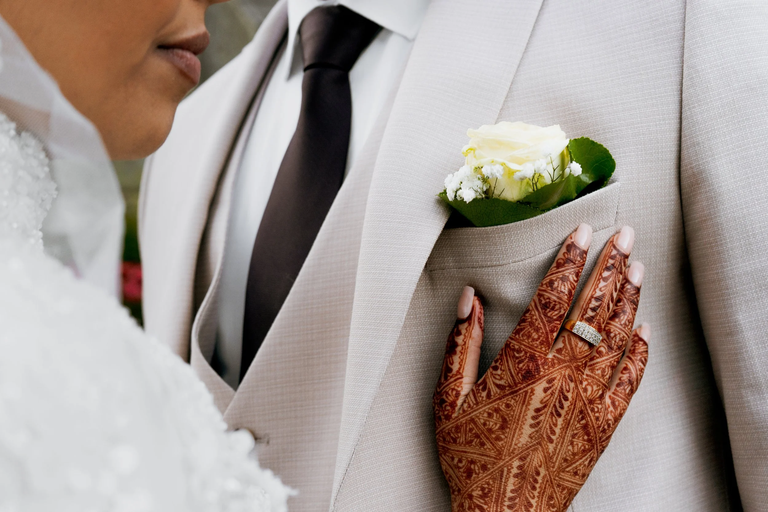photographe mariage lyon Détails d'un mariage montrant un homme en costume beige avec une fleur blanche en boutonnière, une femme avec un doigt tatoué et une bague, et une main posée sur la veste de l'homme avec un tatouage henné.