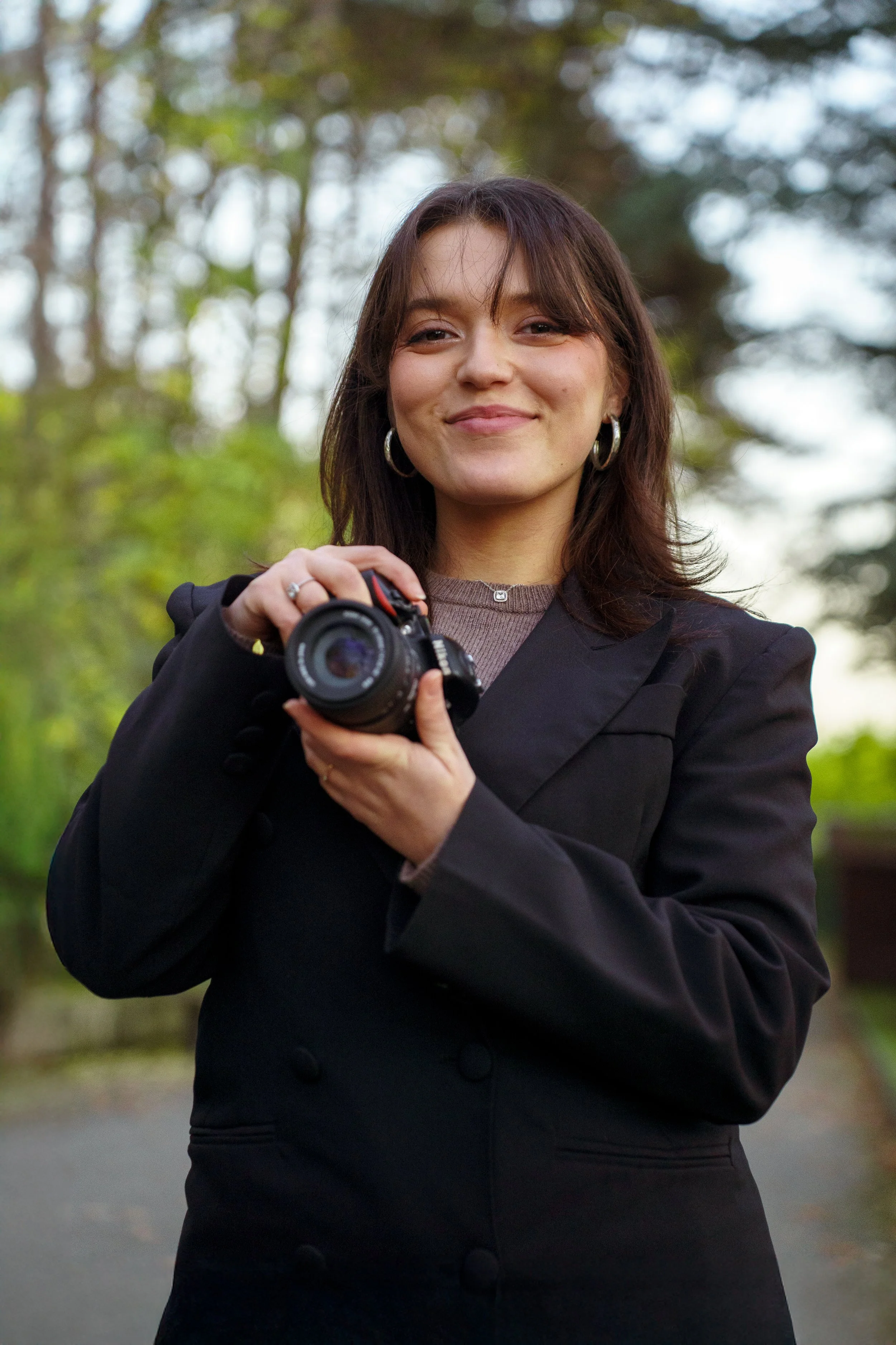 Femme avec un appareil photo, portant une veste noire, debout dans un environnement naturel avec des arbres en arrière-plan.