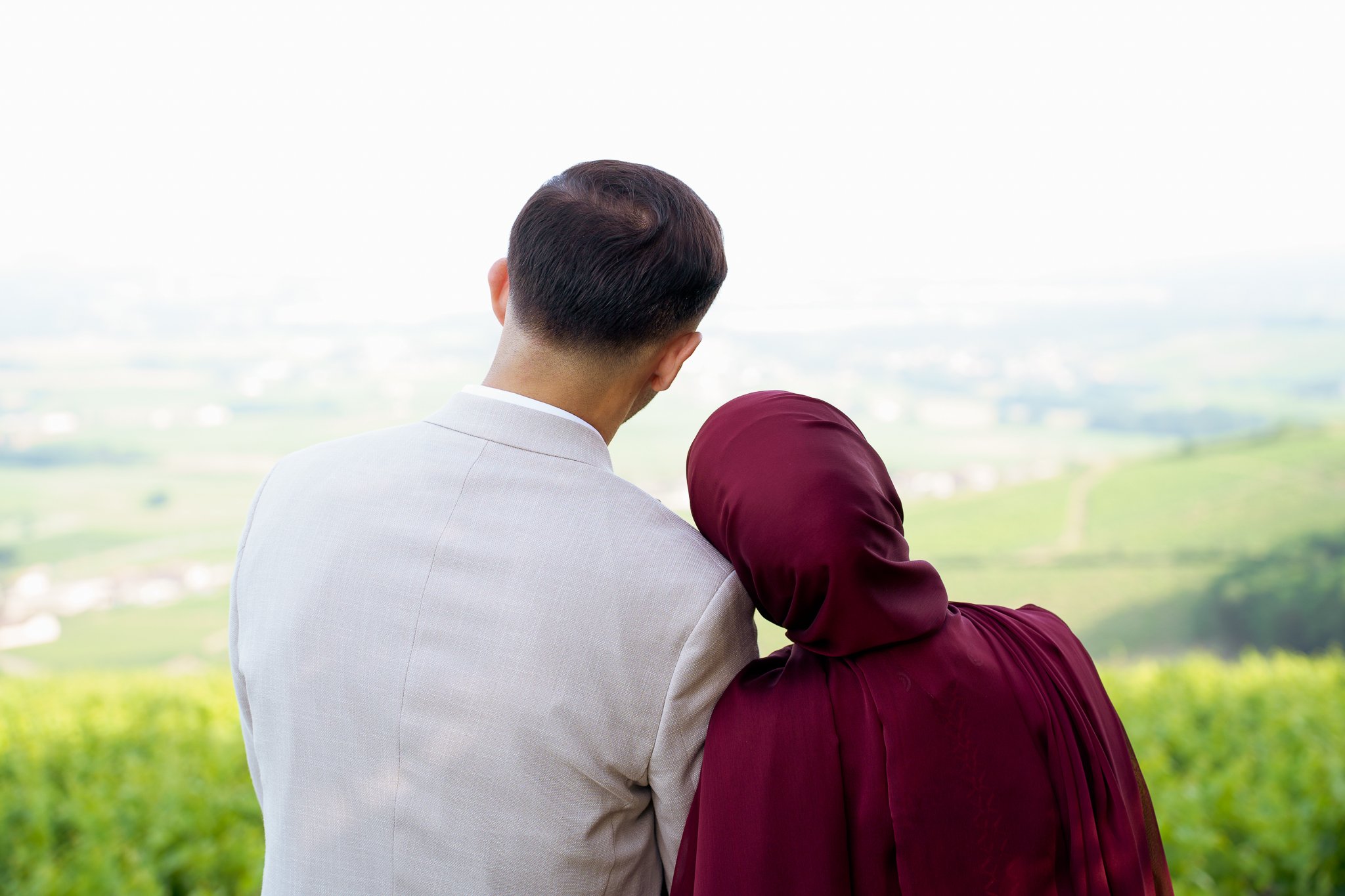 Un couple regarde un paysage verdoyant à l'horizon, l'homme porte un costume clair et la femme porte un hijab bordeaux.