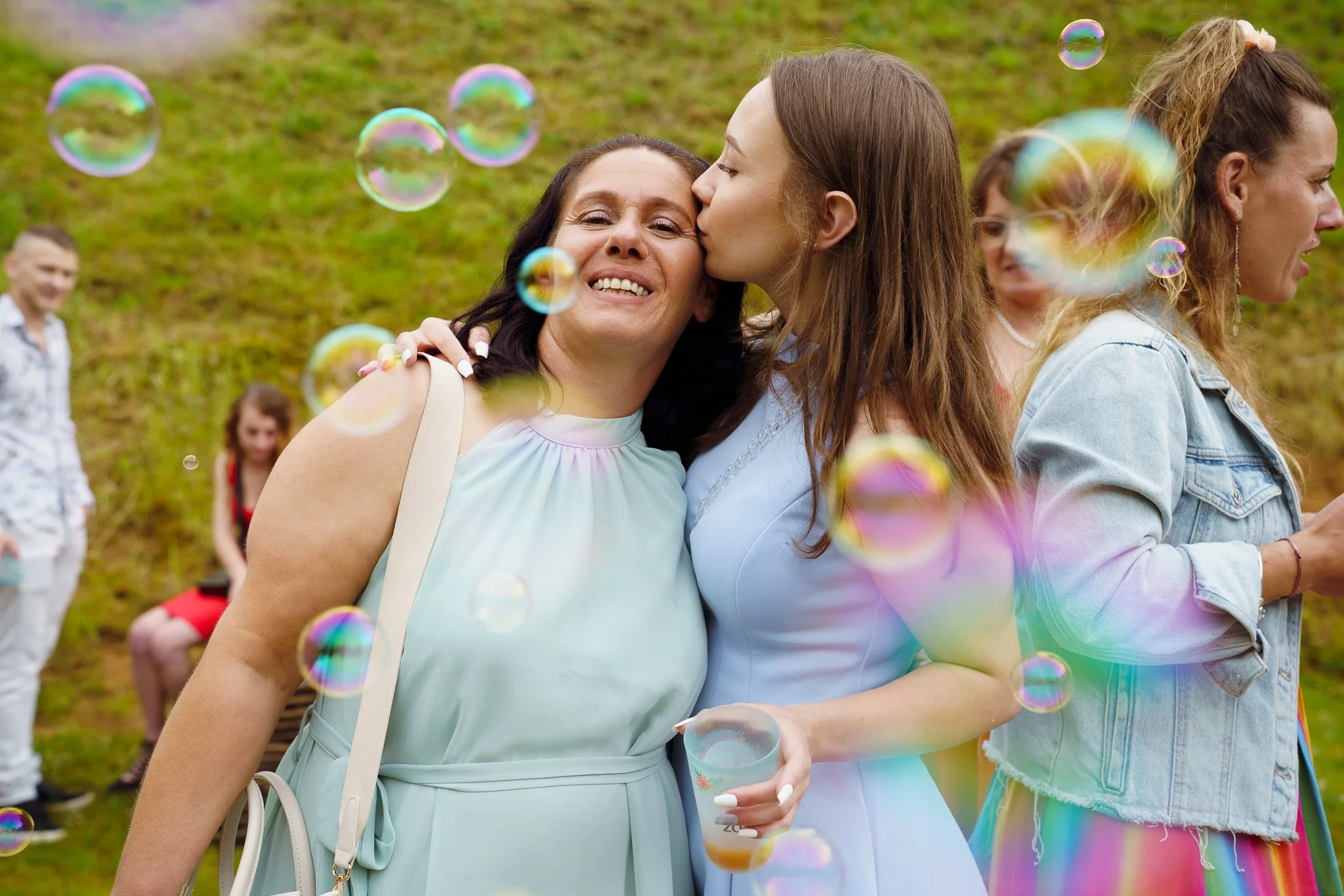 Cinq femmes et deux hommes lors d'une fête en plein air, avec des bulles dans l'air. Deux femmes au centre s'embrassent, l'une leur donnant un baiser sur la joue, dans un environnement verdoyant.