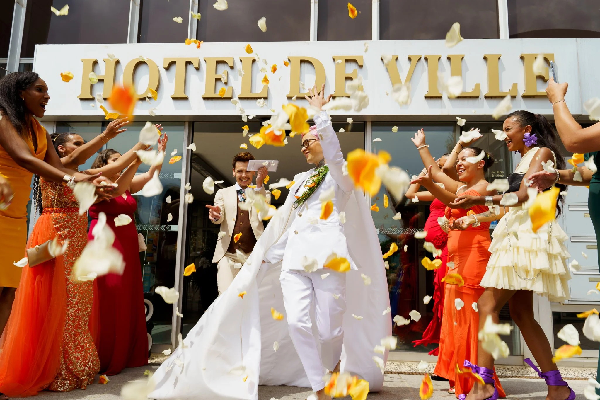 Un groupe de personnes célèbrent un mariage devant un hôtel, avec des fleurs tombant du ciel, et les mariés souriants au centre.
