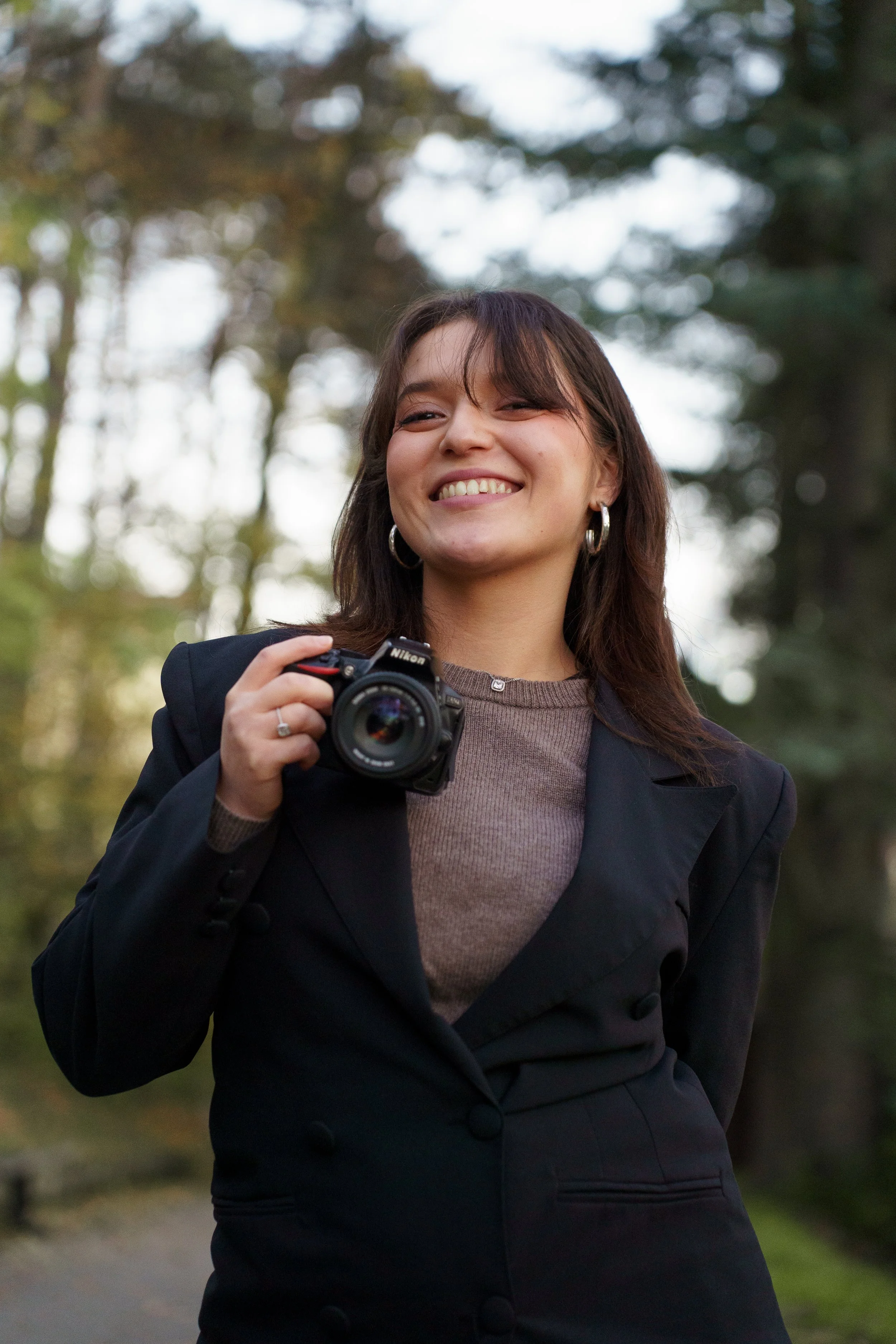 Une femme souriante tenant un appareil photo Nikon, debout dans un parc avec des arbres en arrière-plan.