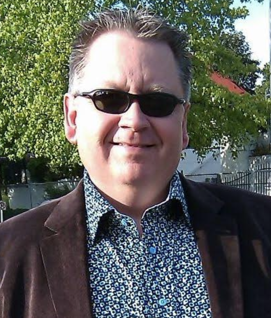 A man wearing sunglasses, a patterned shirt, and a brown jacket standing outdoors with trees and a building in the background.