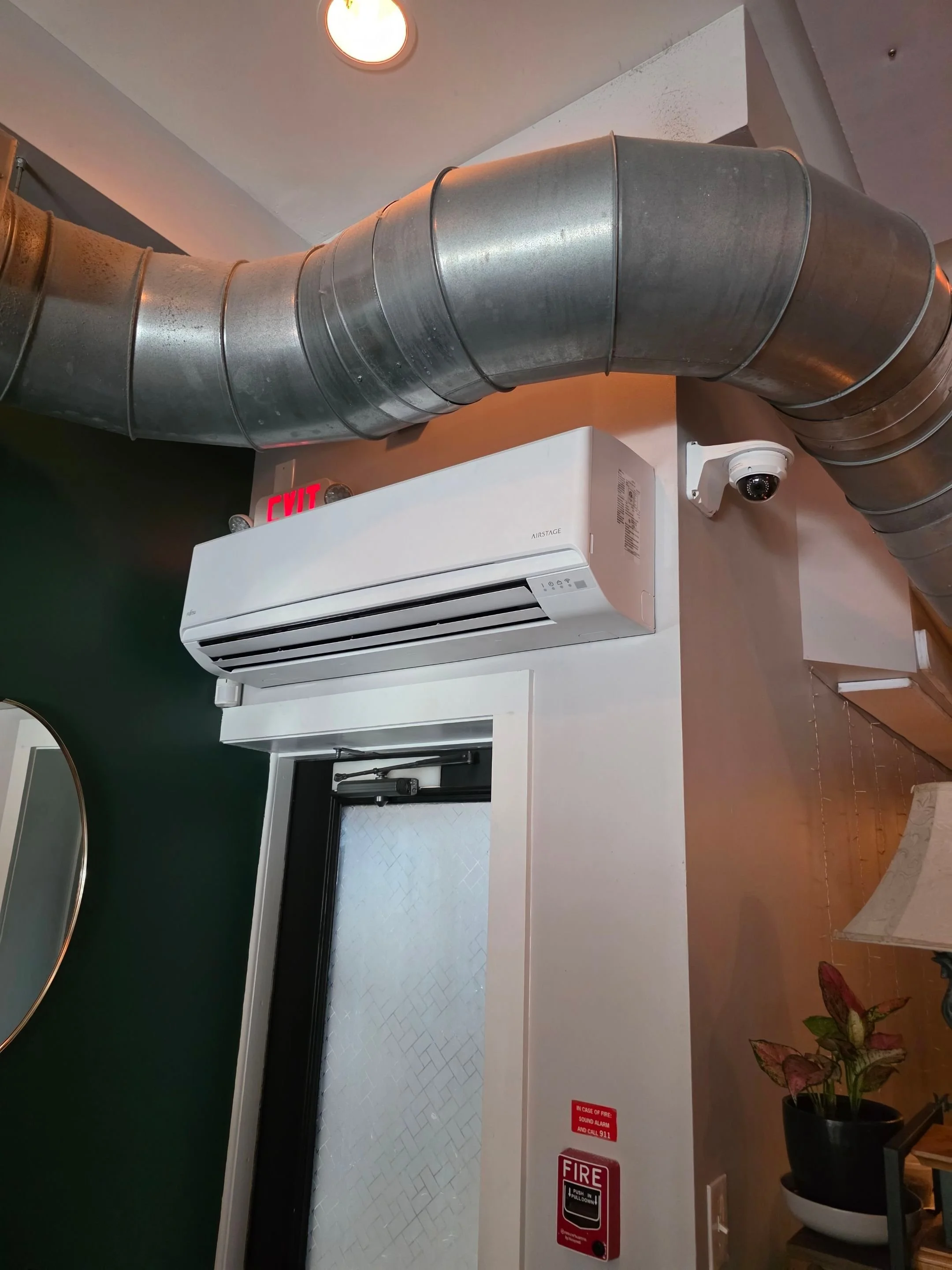 An air conditioning unit mounted on a wall, with large metallic ventilation ducts above it. A security camera is nearby, and signs indicating a fire alarm and emergency exit are visible.