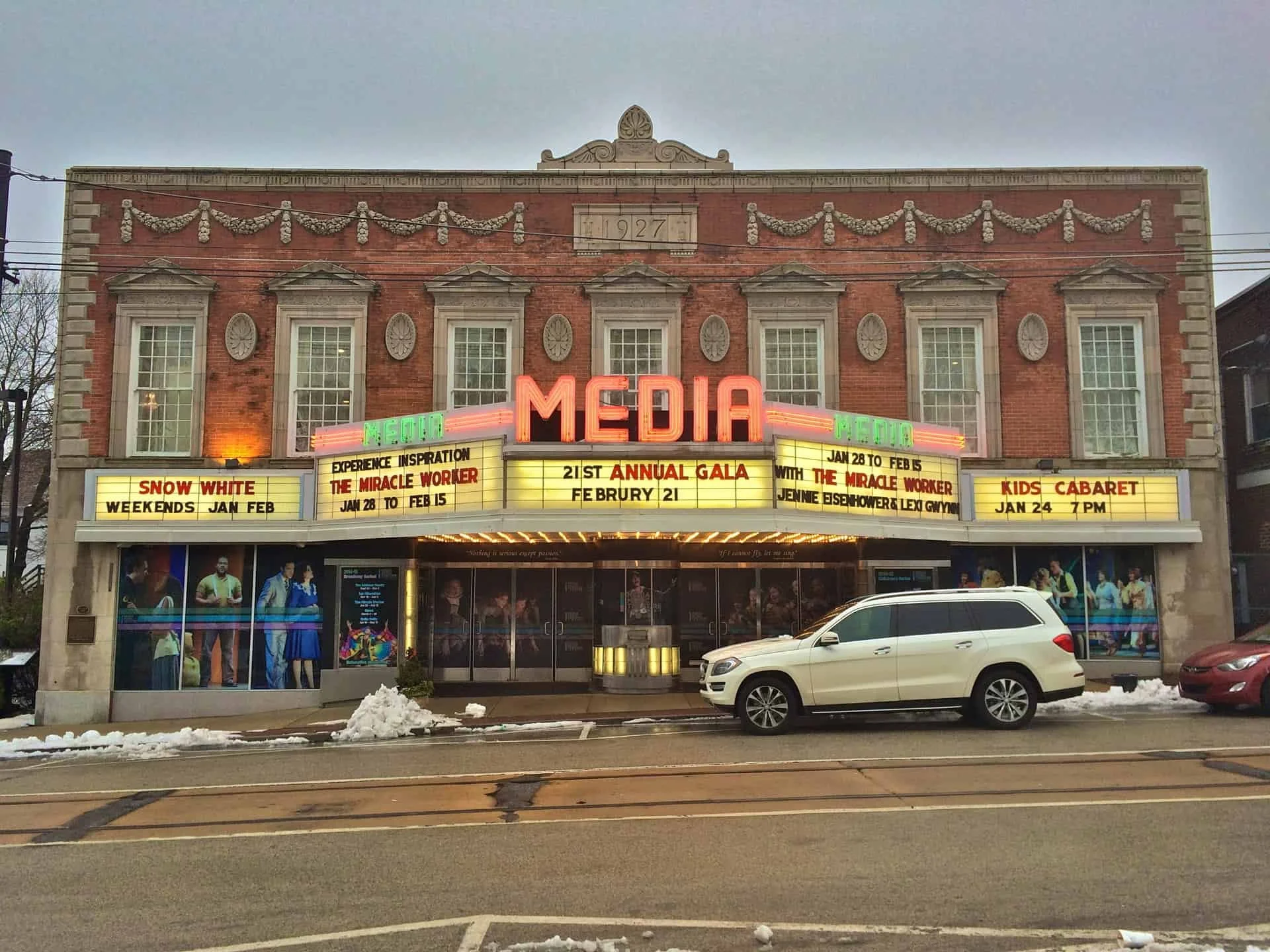 The exterior of a historic theater building with a marquee advertising various events, including a snow white show, a 21st annual gala, and a kids' cabaret. The building has a brick facade with decorative elements, large windows, and a neon sign that reads 'MEDIA'.