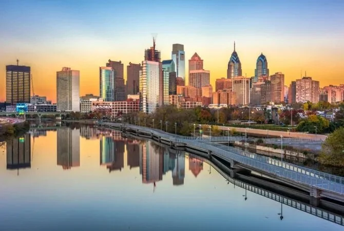 Philadelphia skyline at sunset with buildings reflecting on the river in foreground.