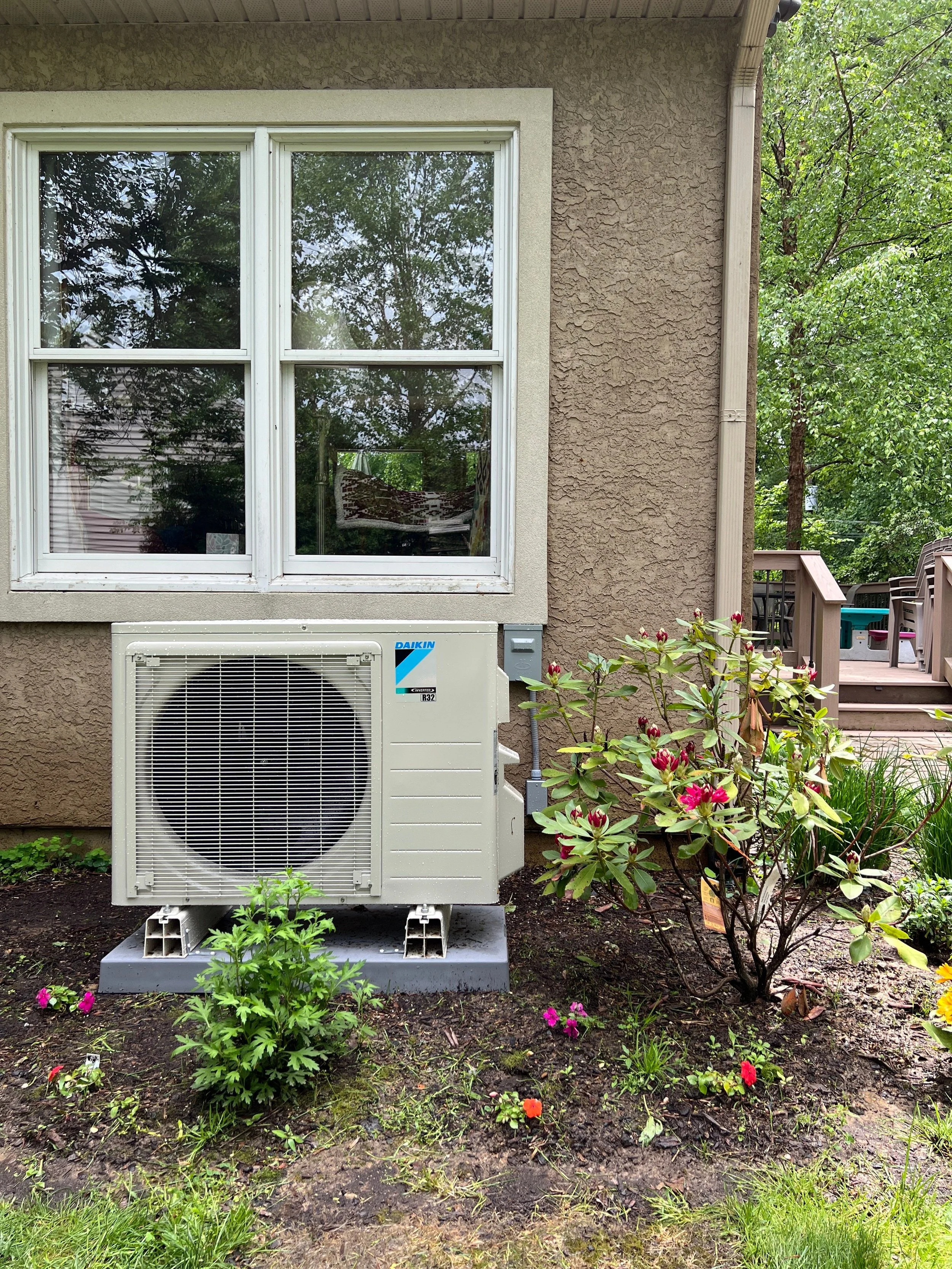 An air conditioning unit installed outside a house next to a garden with a flowering shrub and small colorful flowers, with a dirty wooden deck and a sliding door in the background.