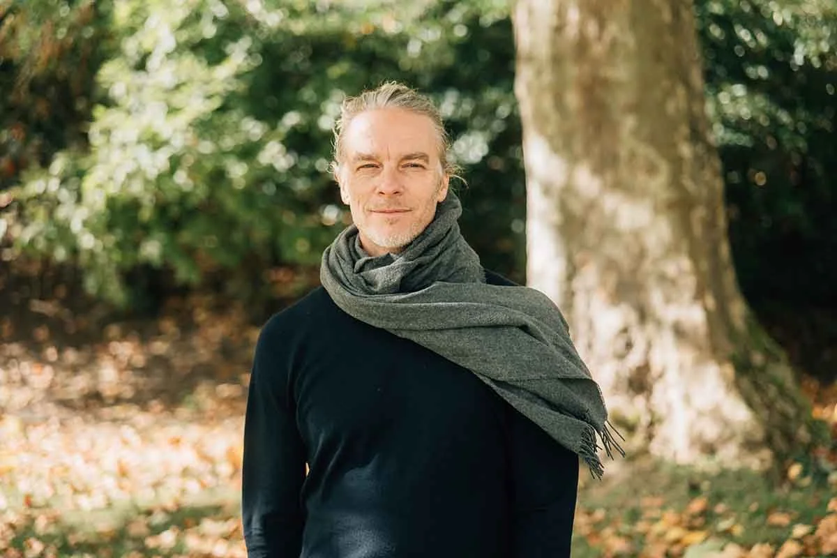 Ein Mann steht im Wald, trägt einen schwarzen Pullover und einen grauen Schal, und hat lange Haare, die zu einem Pferdeschwanz gebunden sind.