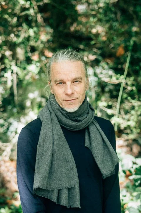 Ein älterer Mann mit grauem Haar und Bart steht im Wald, trägt einen schwarzen Pullover und einen grauen Schal.