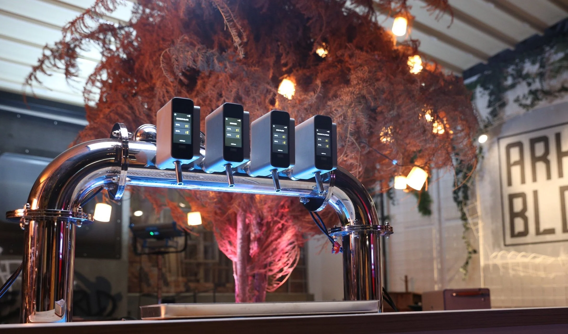 A stainless steel draft beer tap with five digital display screens mounted on top, in front of an autumn-themed background with a decorative reddish-orange tree and hanging lanterns, in an indoor setting with muted lighting.