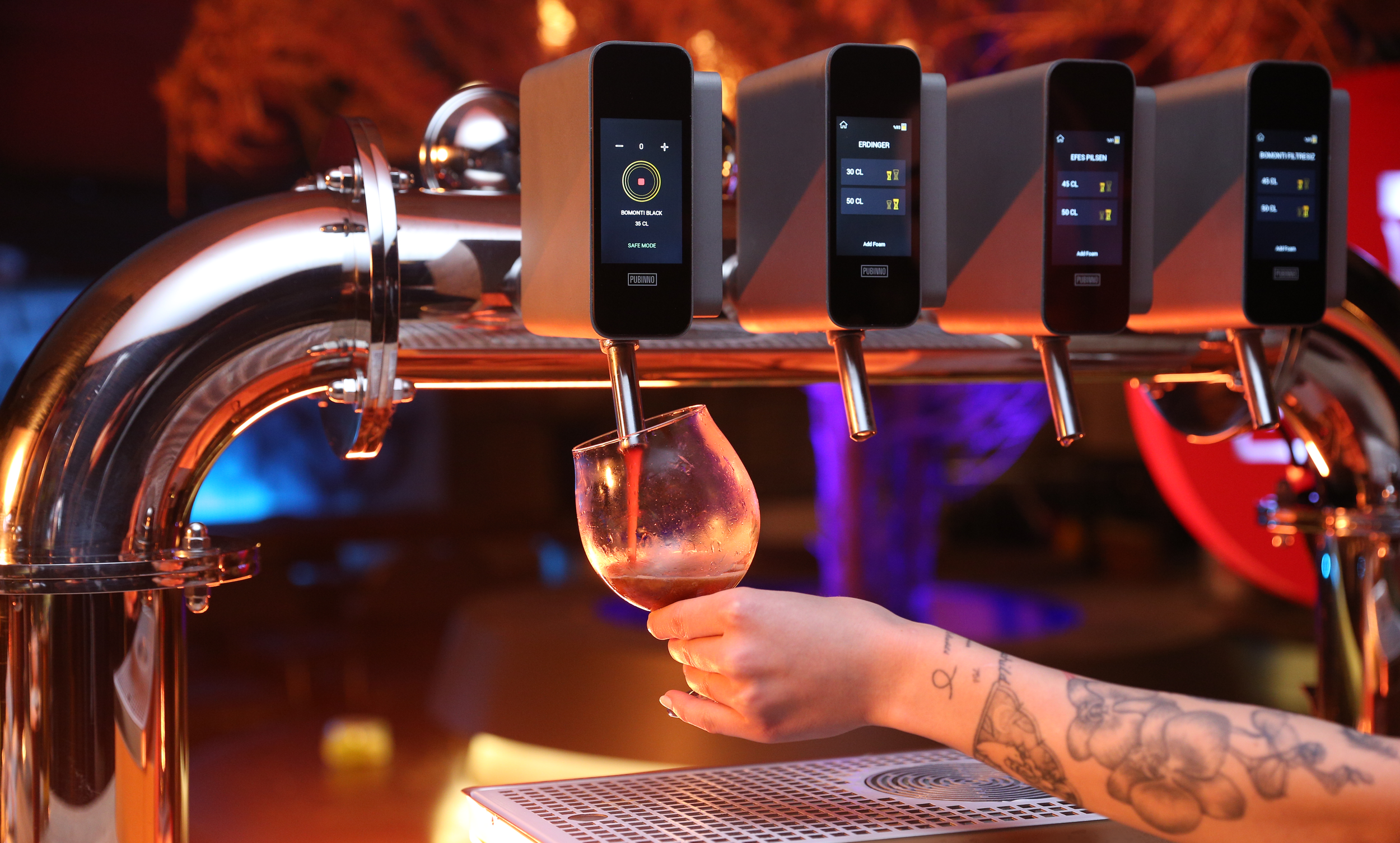 A bartender with tattooed arm holding a glass under a tap in a bar, with multiple electronic devices attached above the tap.