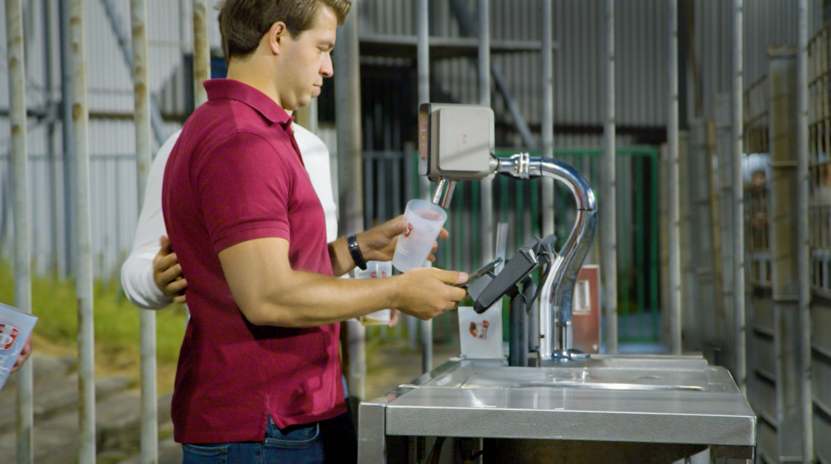 A man in a red polo shirt standing at a public water fountain with a sanitizer dispenser attached, holding a paper cup and using his phone, while another person in a white shirt stands behind him with their hand on his shoulder.