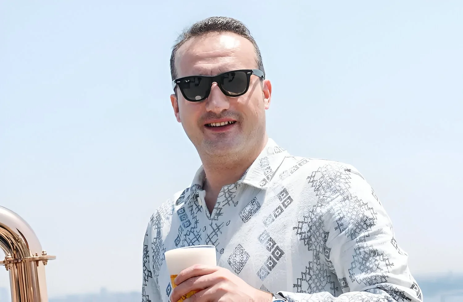 A man wearing black sunglasses and a patterned white shirt, holding a drink, smiling outdoors with a clear sky in the background.