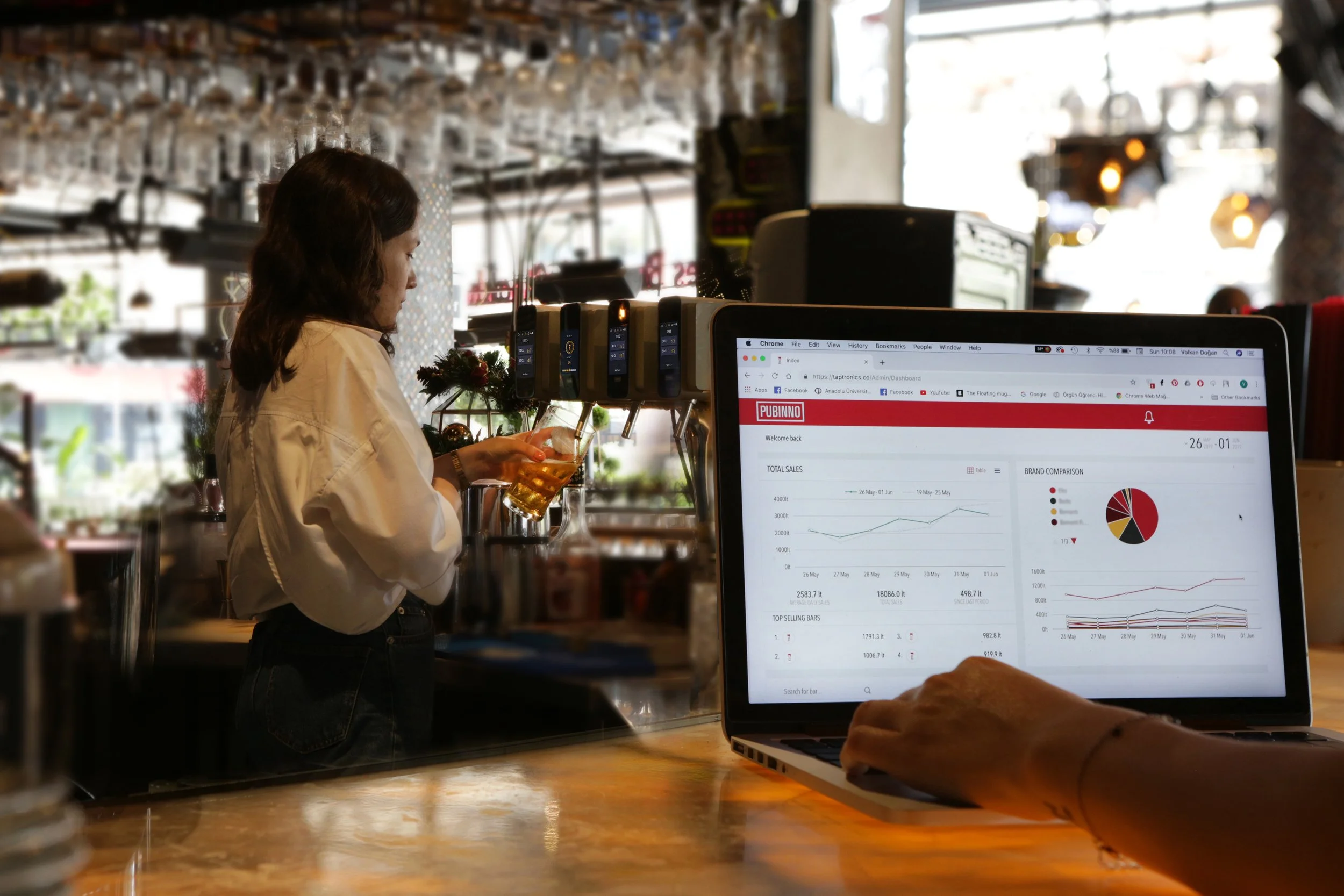 A woman ordering drinks at a bar, with a person working on a laptop displaying sales data on a screen in the foreground.