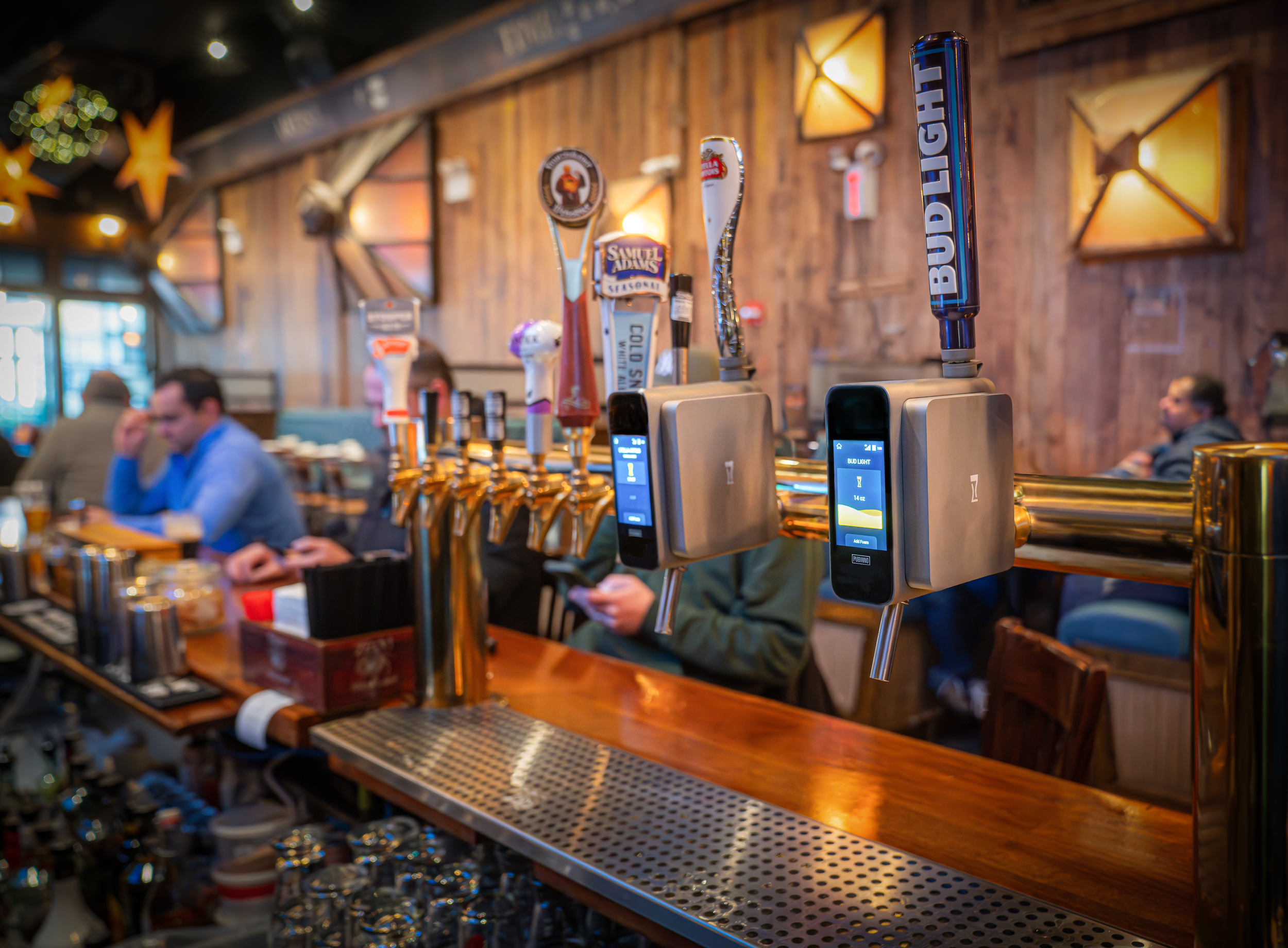 Close-up of beer tap handles at a bar, with patrons in the background. The bar has a wooden interior with warm lighting.