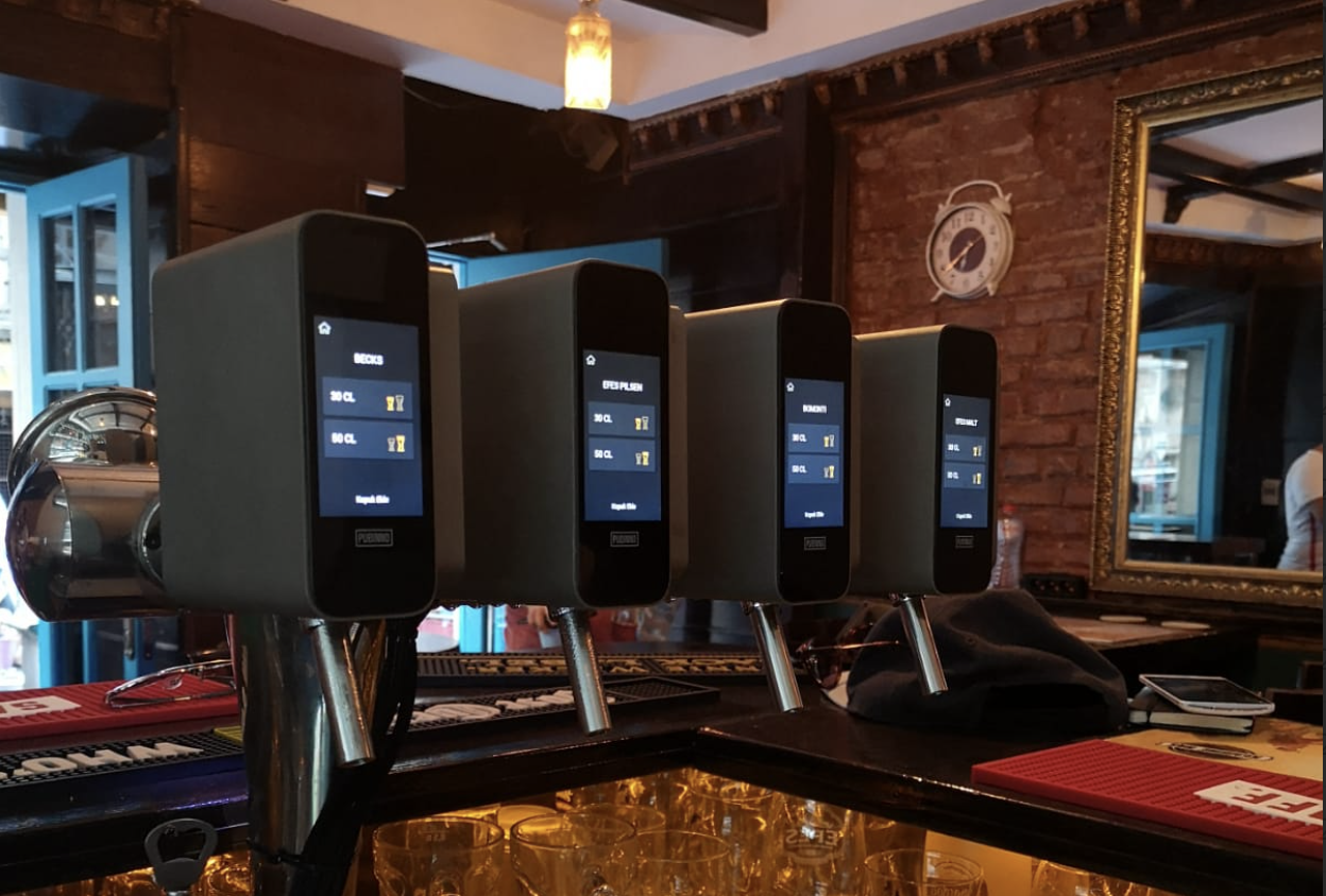 Four beer dispensing machines with digital screens on a bar counter, with glasses underneath and a brick wall with a clock and a mirror in the background.
