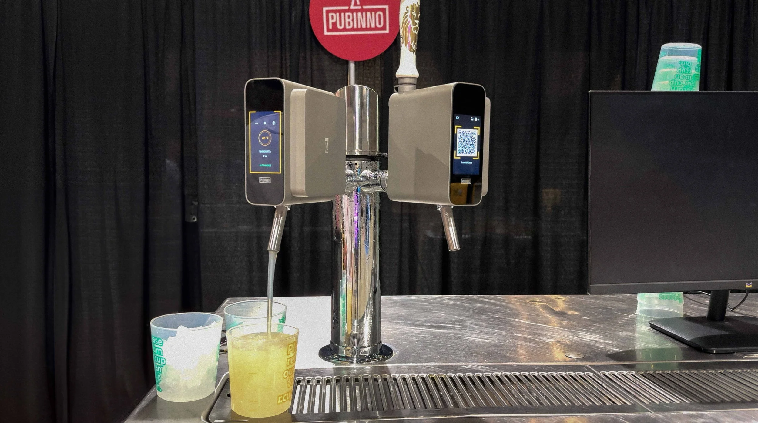 A beer tap system with two digital control panels, dispensing beer into a clear cup on a stainless steel counter, with a computer monitor and black curtain in the background.