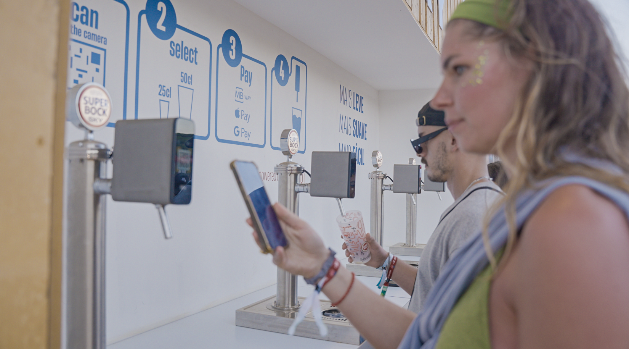 People standing at a self-serve drink station, holding smartphones and cups, with instructional signage on the wall.