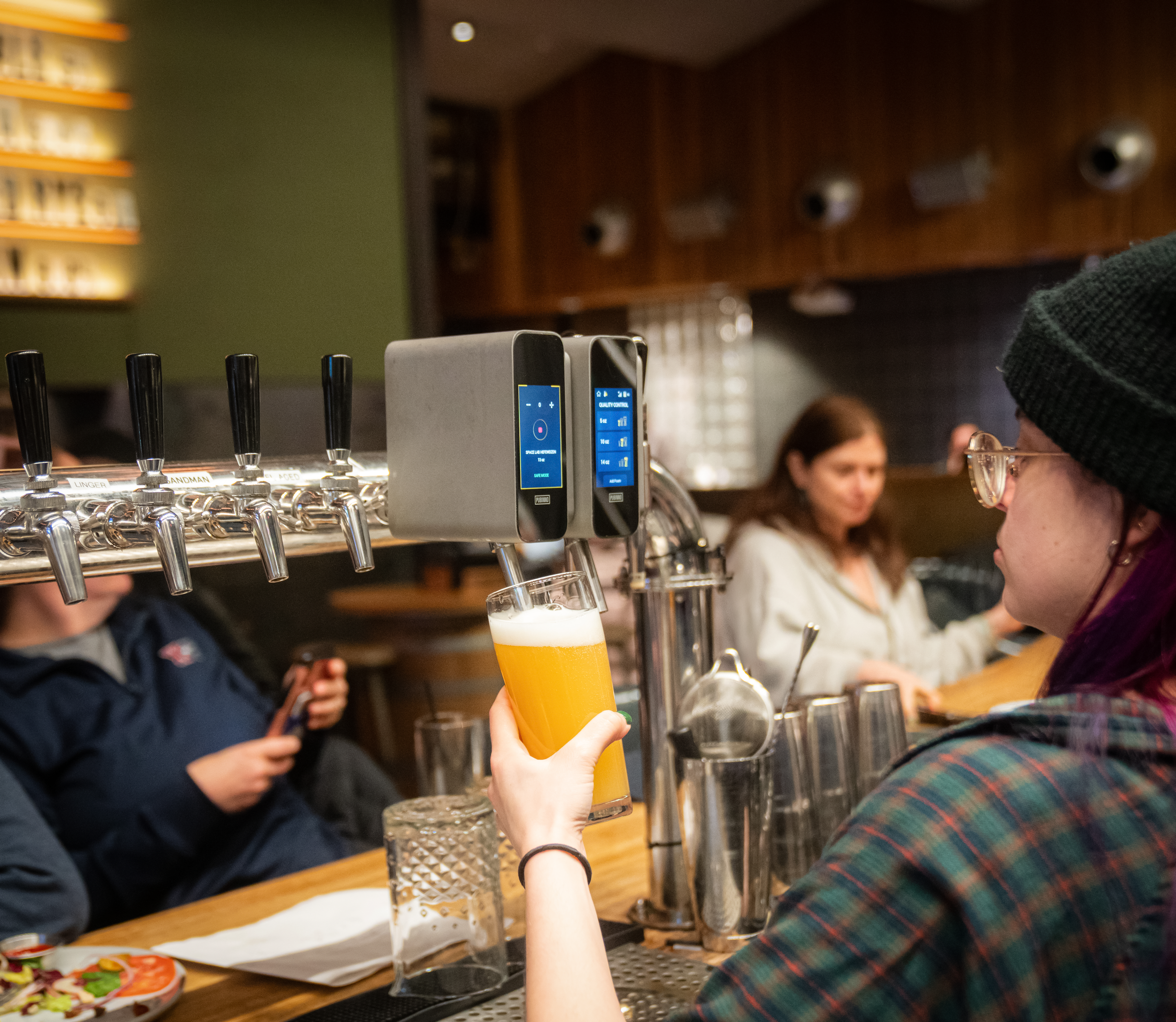 A woman wearing glasses, a black beanie, and a plaid shirt is pouring a beer from a tap at a bar. There are two digital screens above the tap, a glass of beer in her hand, and other bar items on the counter. In the background, there are people sitting and talking.