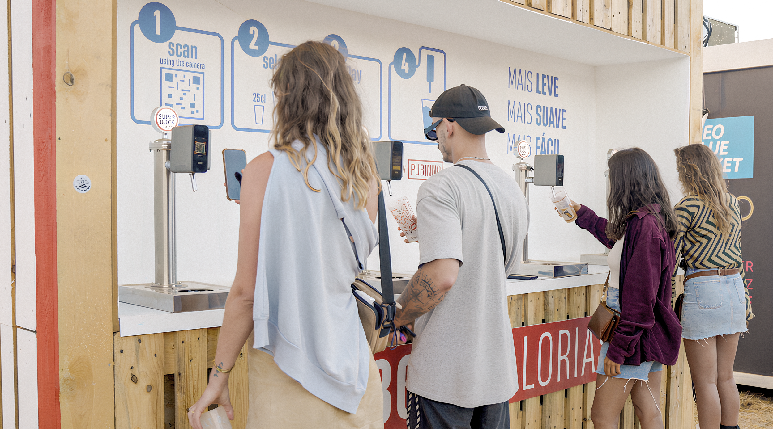 Four people standing in line at a beverage stall, using their phones, one holding a drink, with a sign explaining the ordering process behind them.