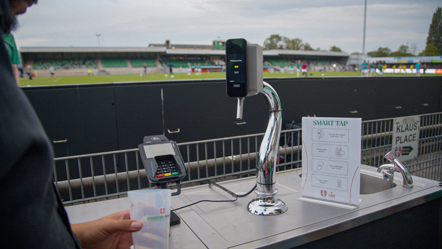 A contactless smart tap payment station with a hand holding a cup in front of it, set up at a sports stadium.