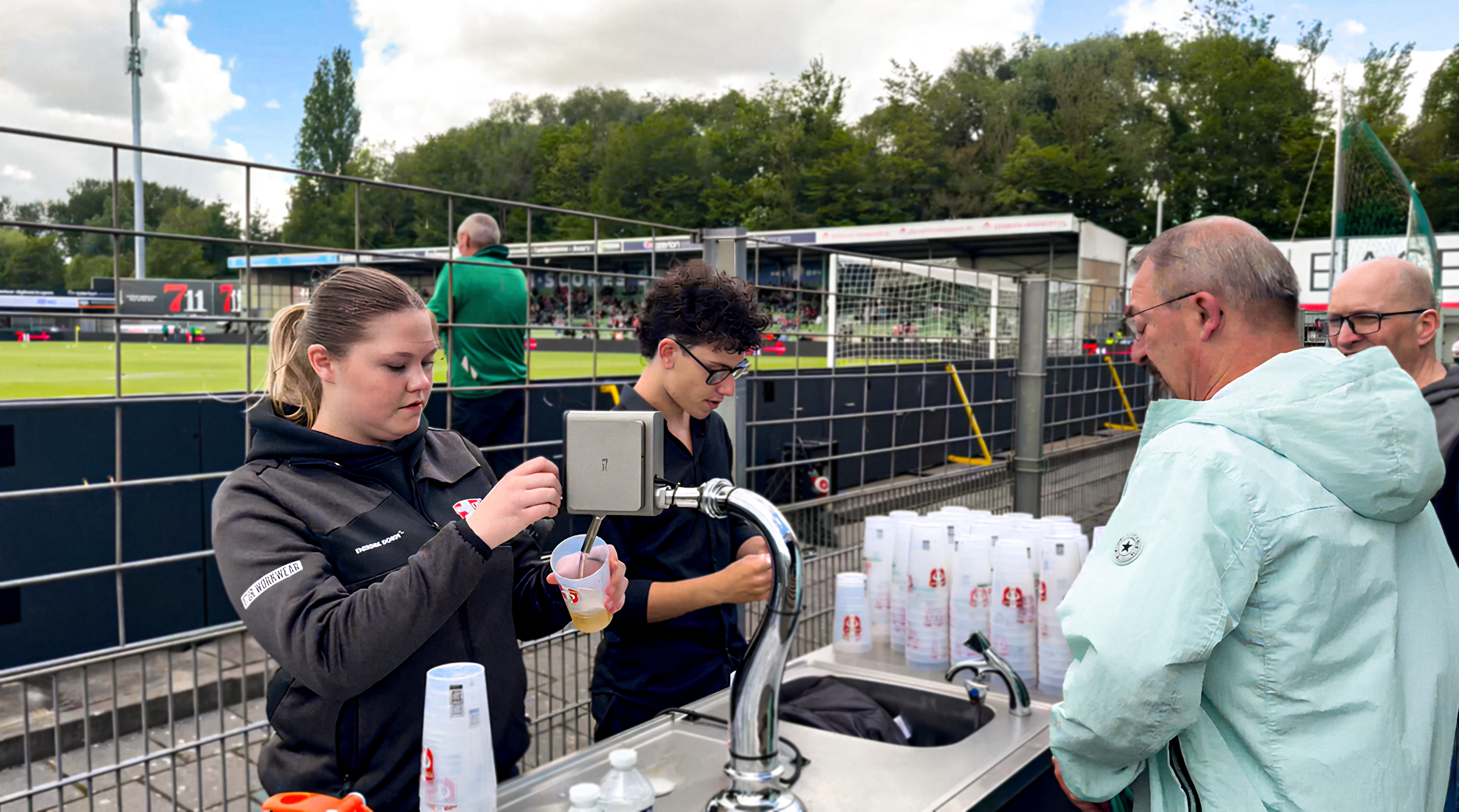 People serving drinks at an outdoor sports stadium concession stand with a soccer field in the background.