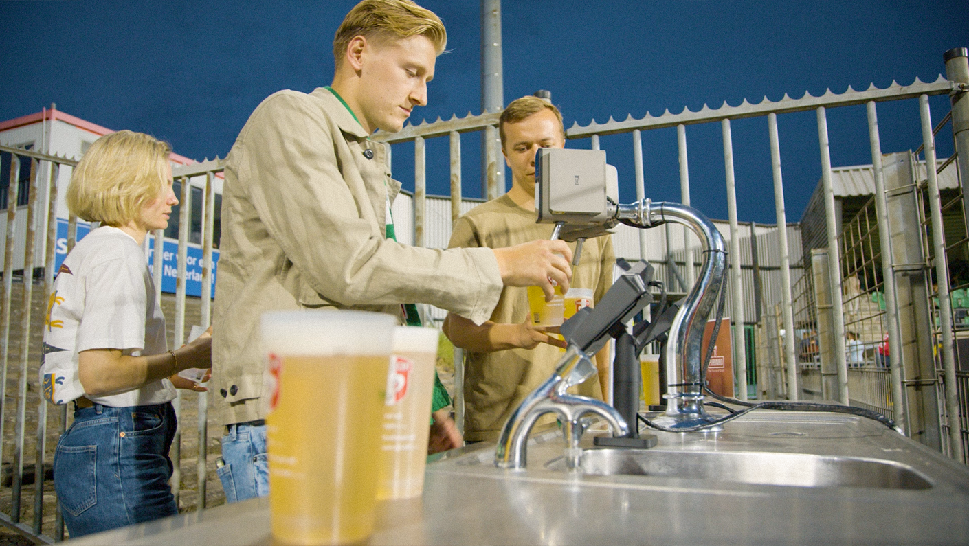 Three people standing in line at a concession stand with a soda fountain, with one person purchasing drinks and two people waiting. It is evening, and the background includes metal fencing and a sign.
