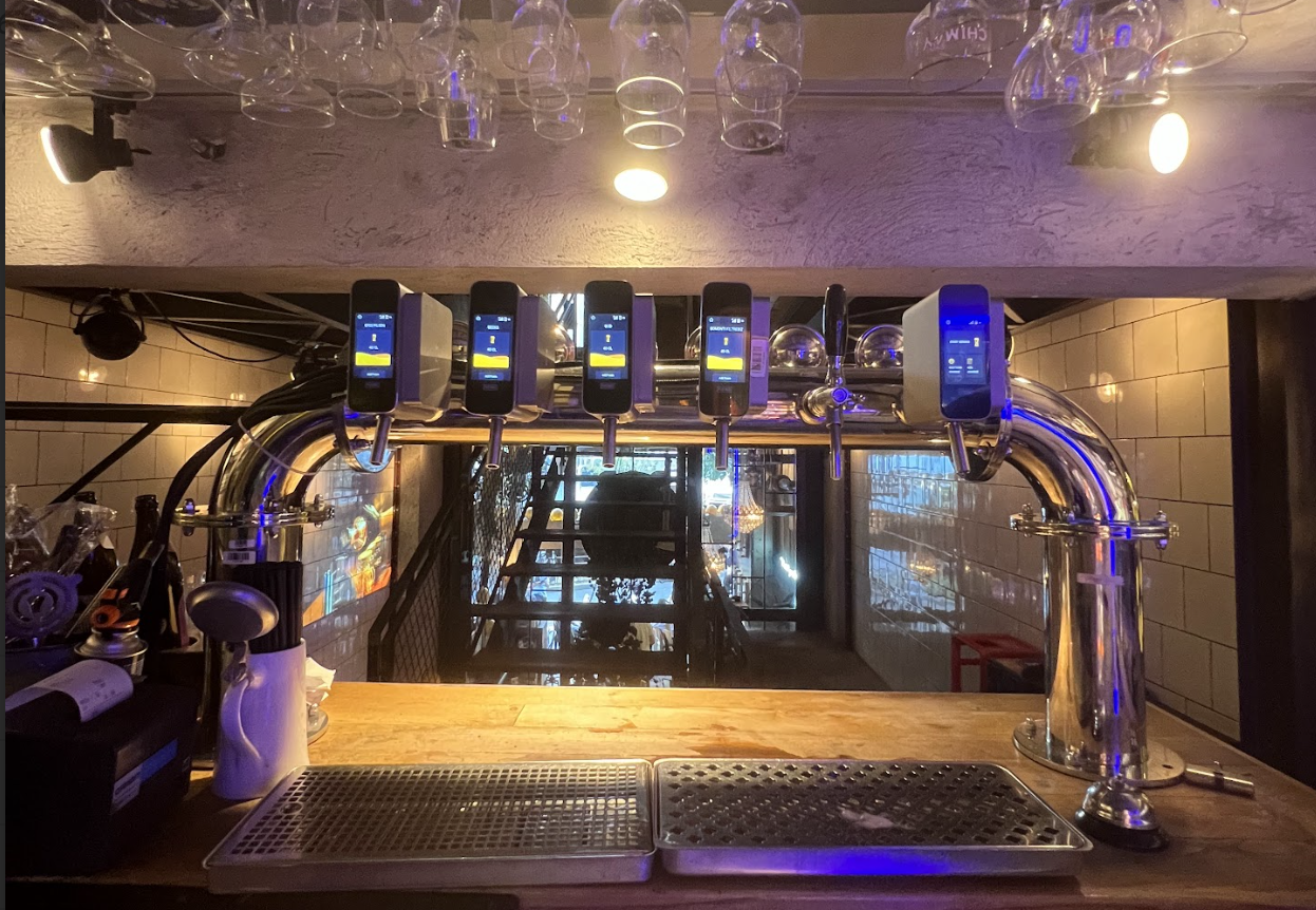 A bar counter with beer taps and glasses hanging above, viewed from behind the bar.