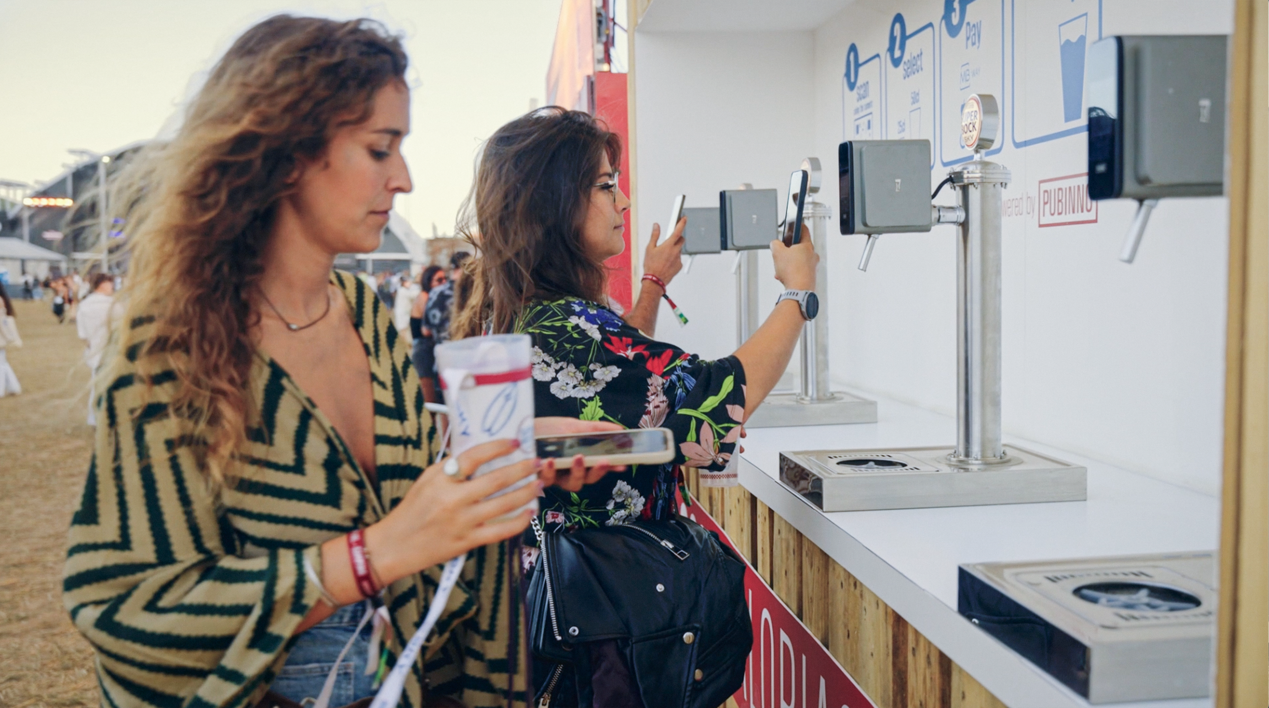 Two women at a ticket or payment kiosk, each holding a smartphone, with one also holding a cup, at an outdoor event or festival.