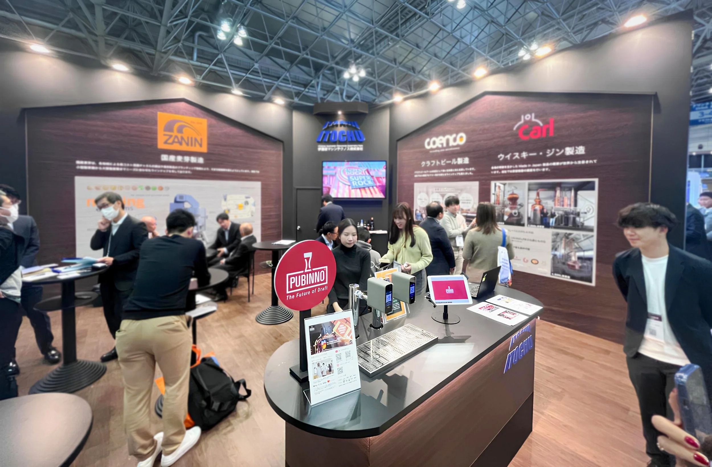 A busy trade show booth with multiple people interacting and discussing, featuring large banners with logos and descriptions of products, a central counter with screens and informational materials, and a modern industrial ceiling.