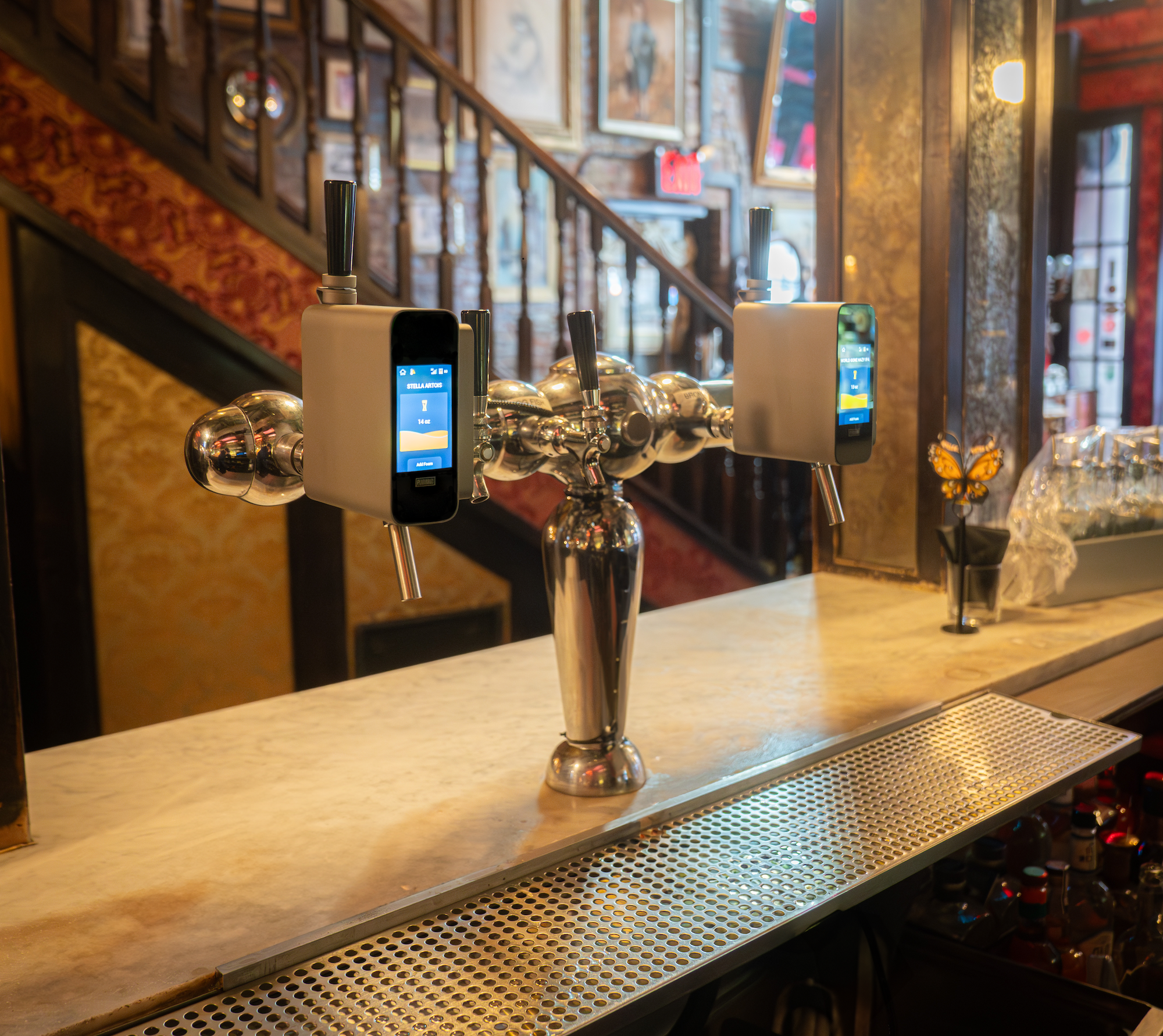 Two self-serve drink dispensers on a bar counter with digital screens and taps, in a warmly decorated restaurant or bar with artwork on the wall, staircase, and a butterfly decoration.
