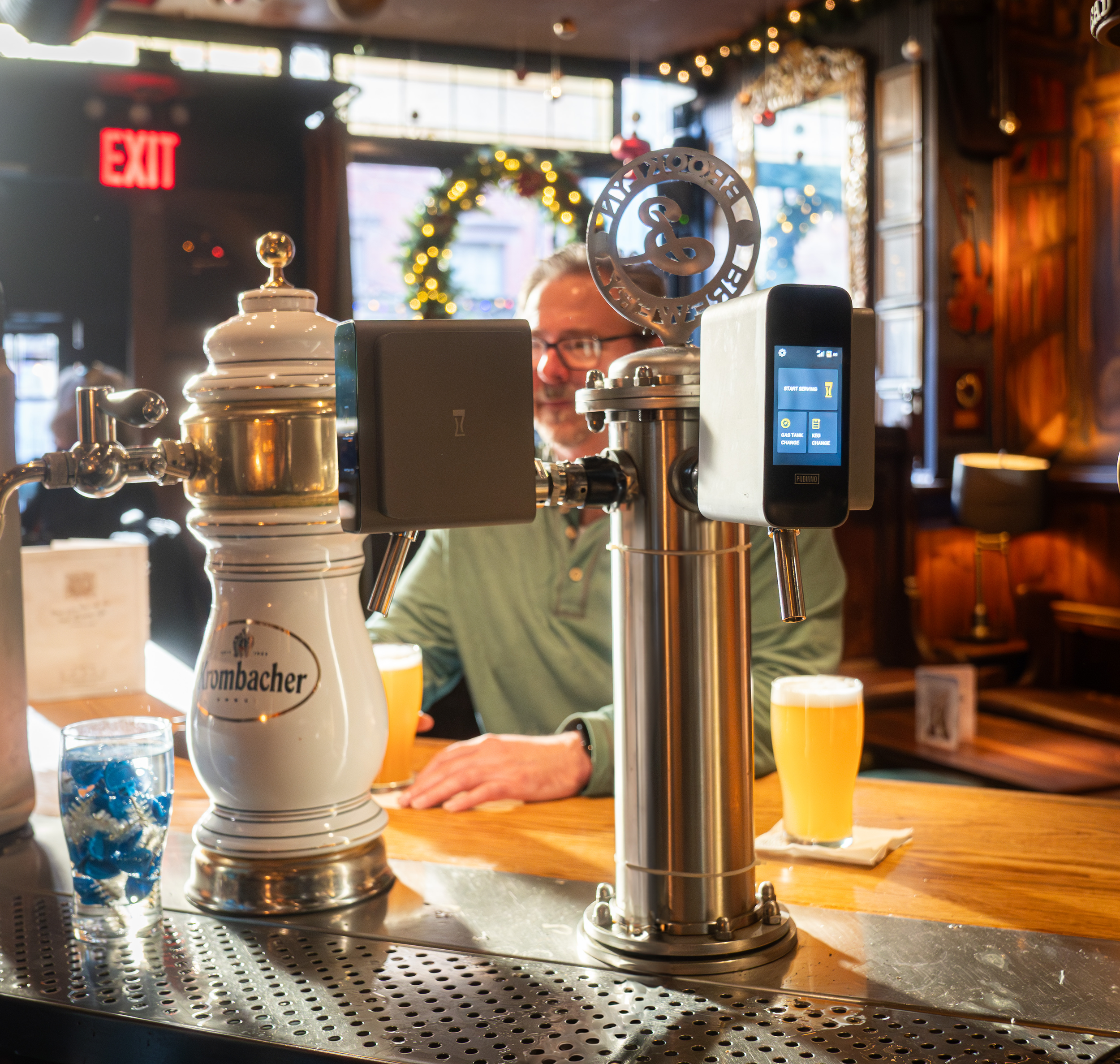 A man sitting at a bar in a restaurant or pub, with a beer tap and a glass of beer in front of him, and a decorative wreath hanging in the background, during the daytime.