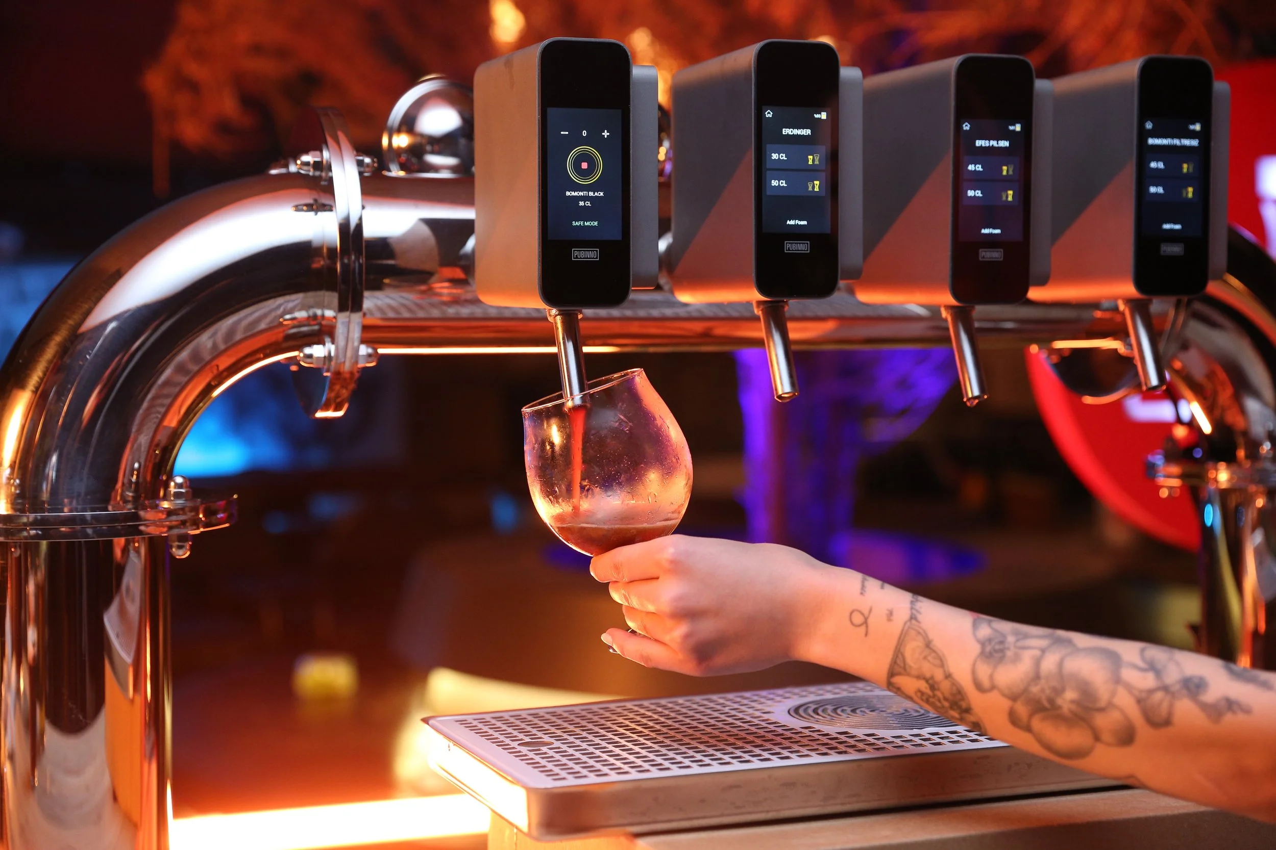 A person with tattoos on their arm holding a wine glass under a tap station with digital drink dispensers in a bar or nightclub setting.