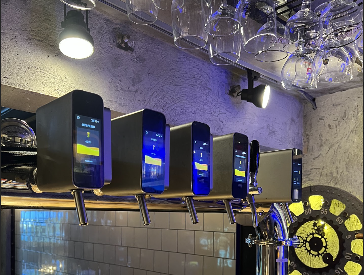 A row of digital beer dispense machines behind a bar, with glasses hanging upside down above and a clock on the wall in the background.