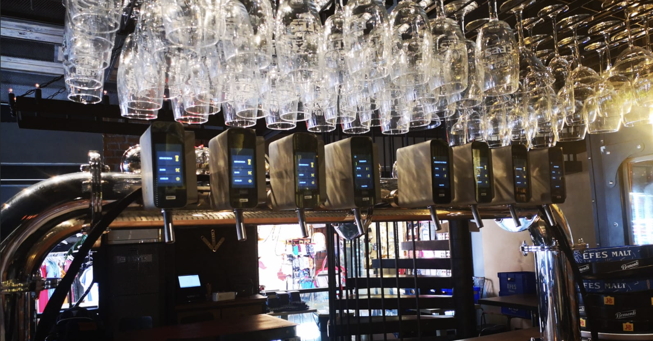 Multiple empty beer glasses hanging upside down above a row of beer taps in a bar or restaurant setting, with sunlight streaming in from the right side.