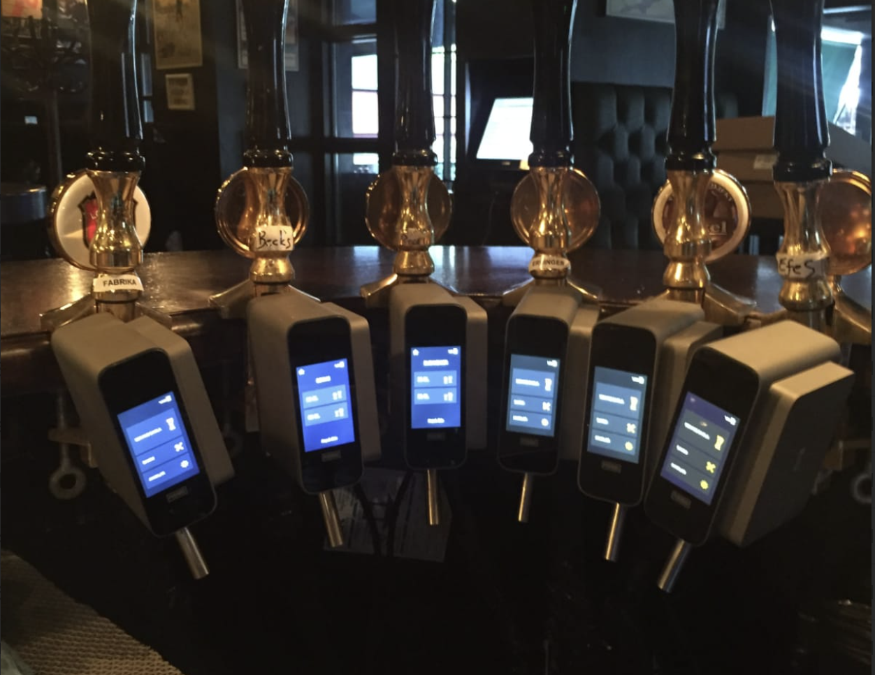 Group of six automatic beer dispensers with digital interfaces in a dark, cozy bar setting.