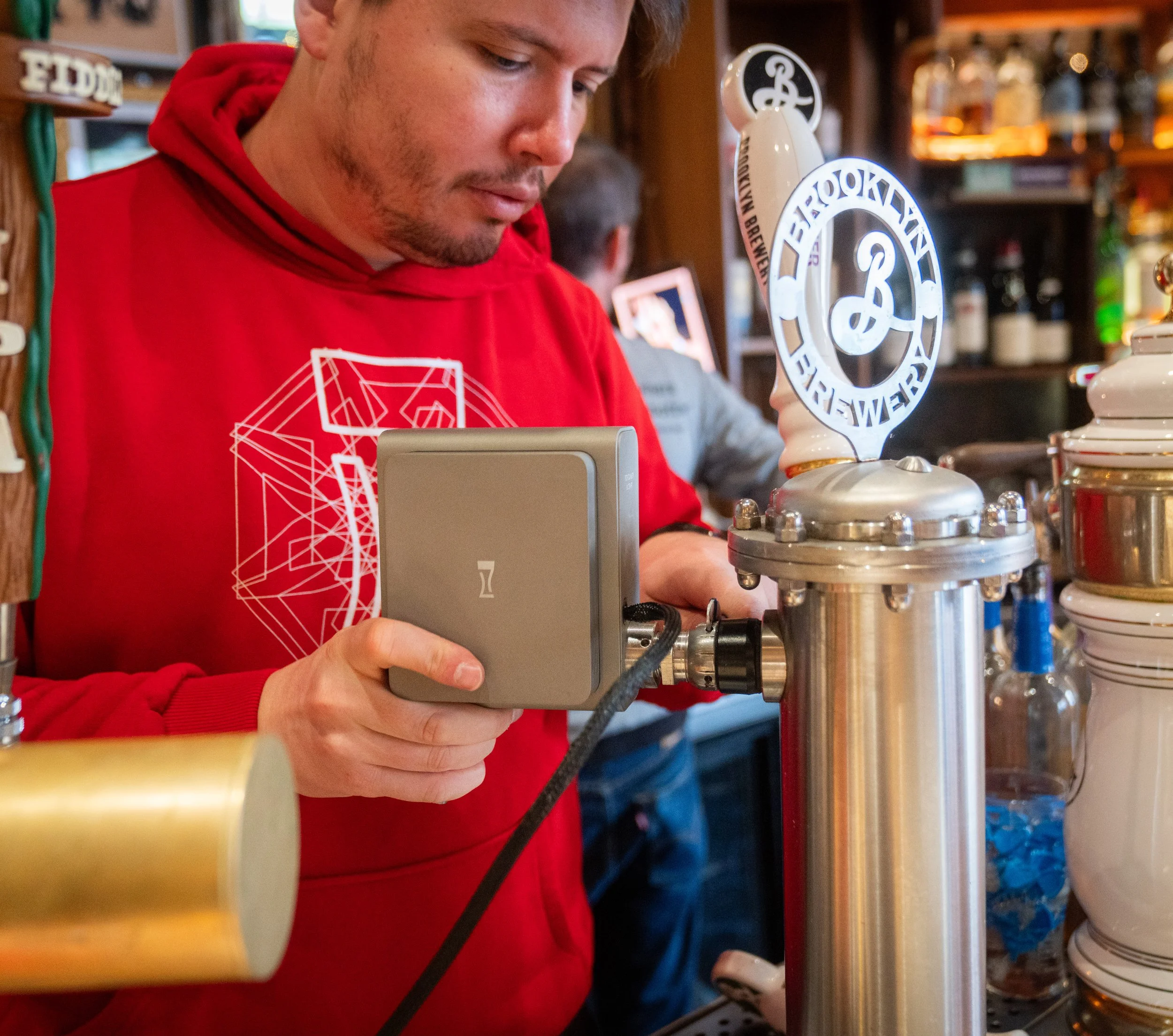 A man in a red hoodie is using a digital device on a beer tap in a bar or brewery, with a background of wooden shelves and bottles.