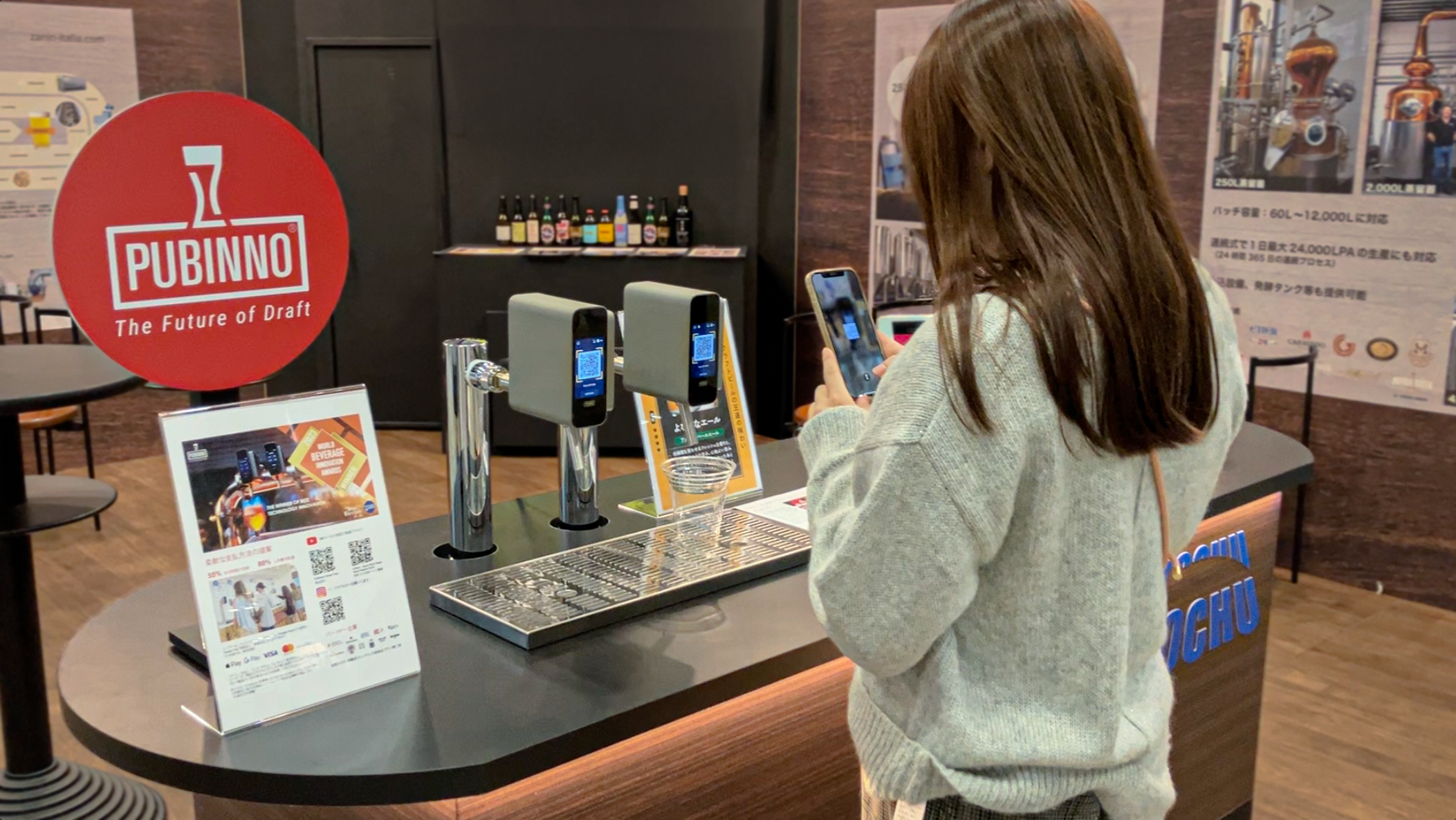 A woman with brown hair wearing a gray sweater and carrying a brown shoulder bag is standing at a bar counter, looking at her smartphone. Behind her, there is a red circular sign with the logo and text 'PUBINNO The Future of Draft'. The counter has t