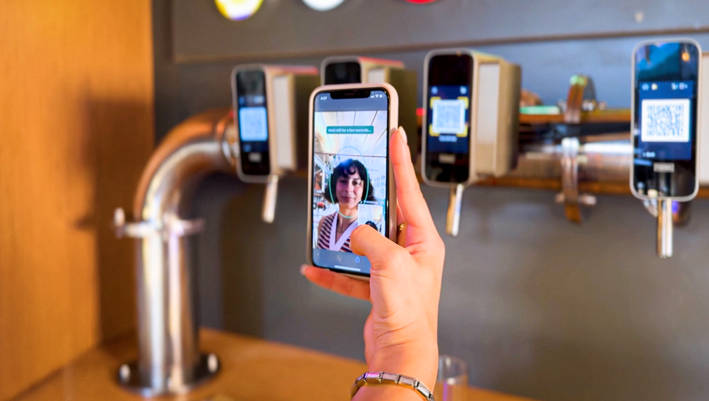 Person taking a photo of a woman with a smartphone at a drink dispenser with multiple smartphones on the dispenser.