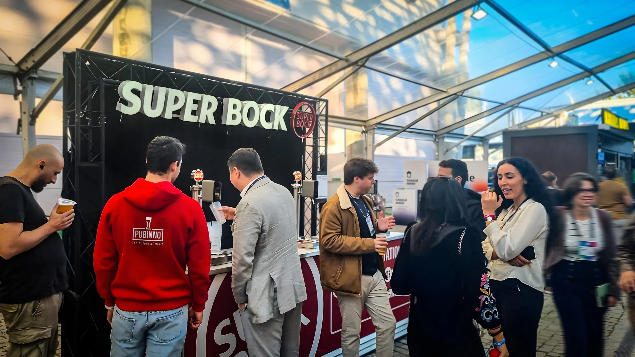People gathered around a beer stand labeled 'Super Bock' under a transparent tent at an event, with some holding beer glasses and engaging in conversation.
