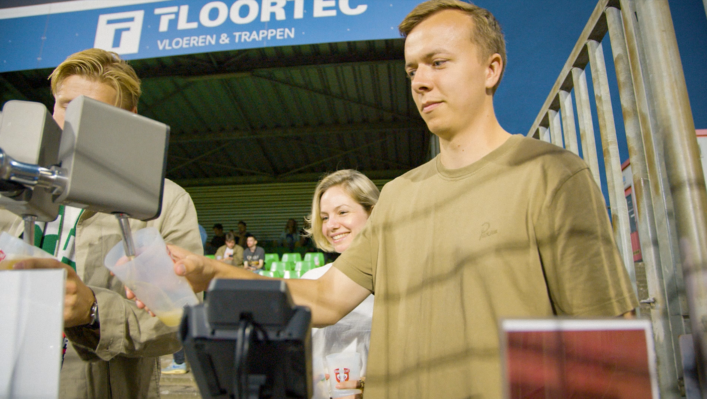 Three people at a checkout counter; two young men, one young woman, smiling, in a large indoor space with seats and a banner overhead.