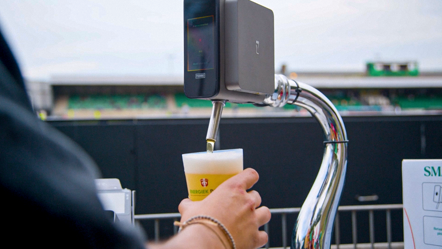 A person holding a plastic cup under a beer tap at a stadium, with a digital payment terminal attached to the tap.