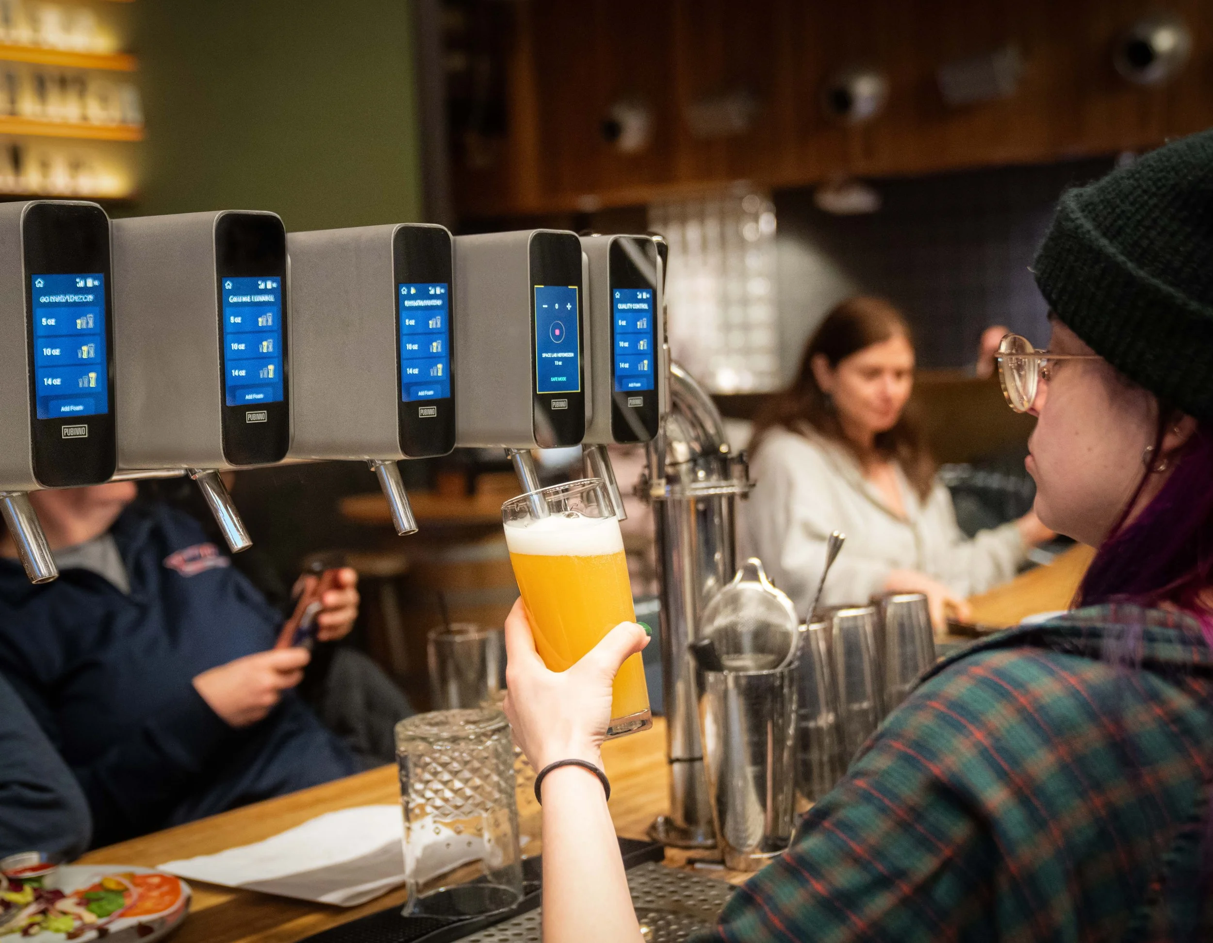 Person with glasses and a beanie pouring a draft beer at a bar, with other people in the background and a row of beer taps.