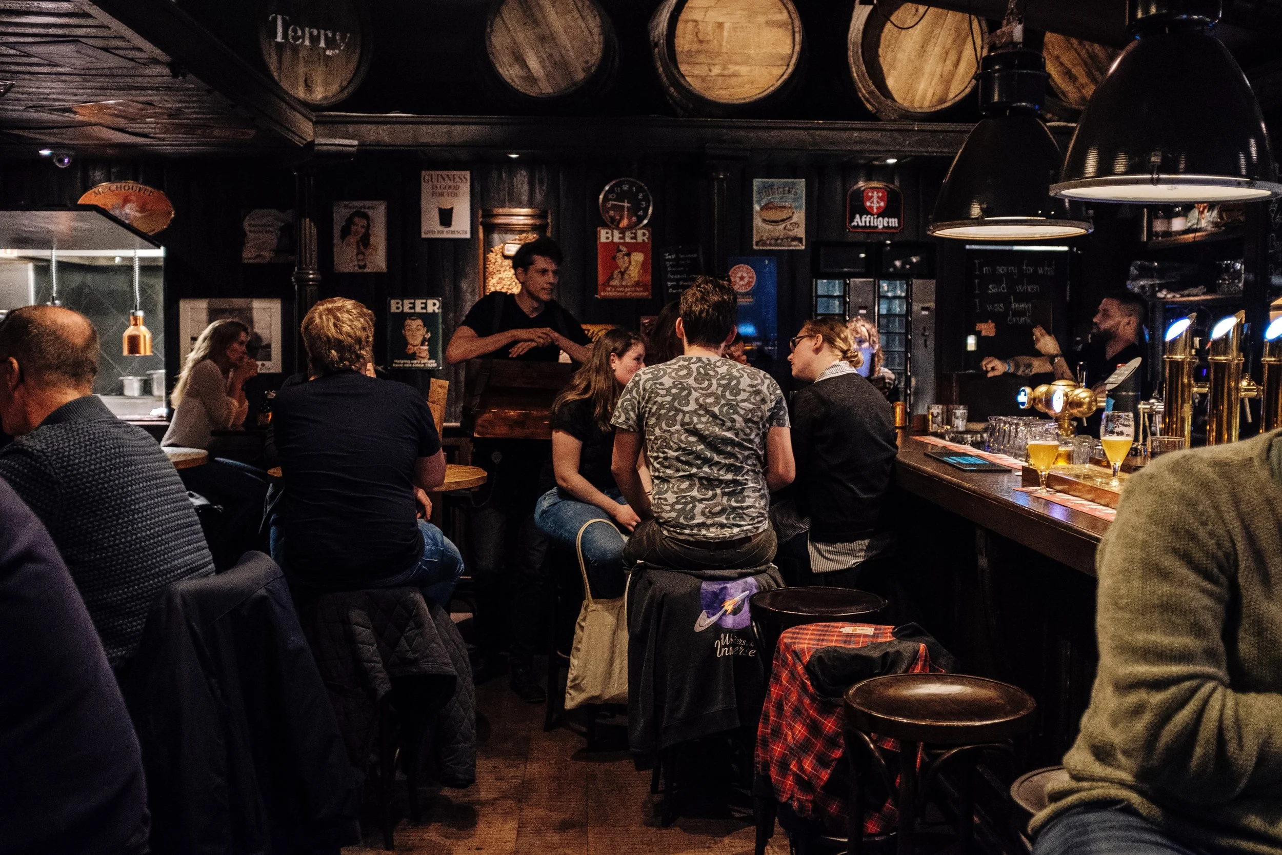 A crowded pub with people sitting at the bar and tables, vintage beer signs on the dark wooden walls, and a bartender serving drinks.
